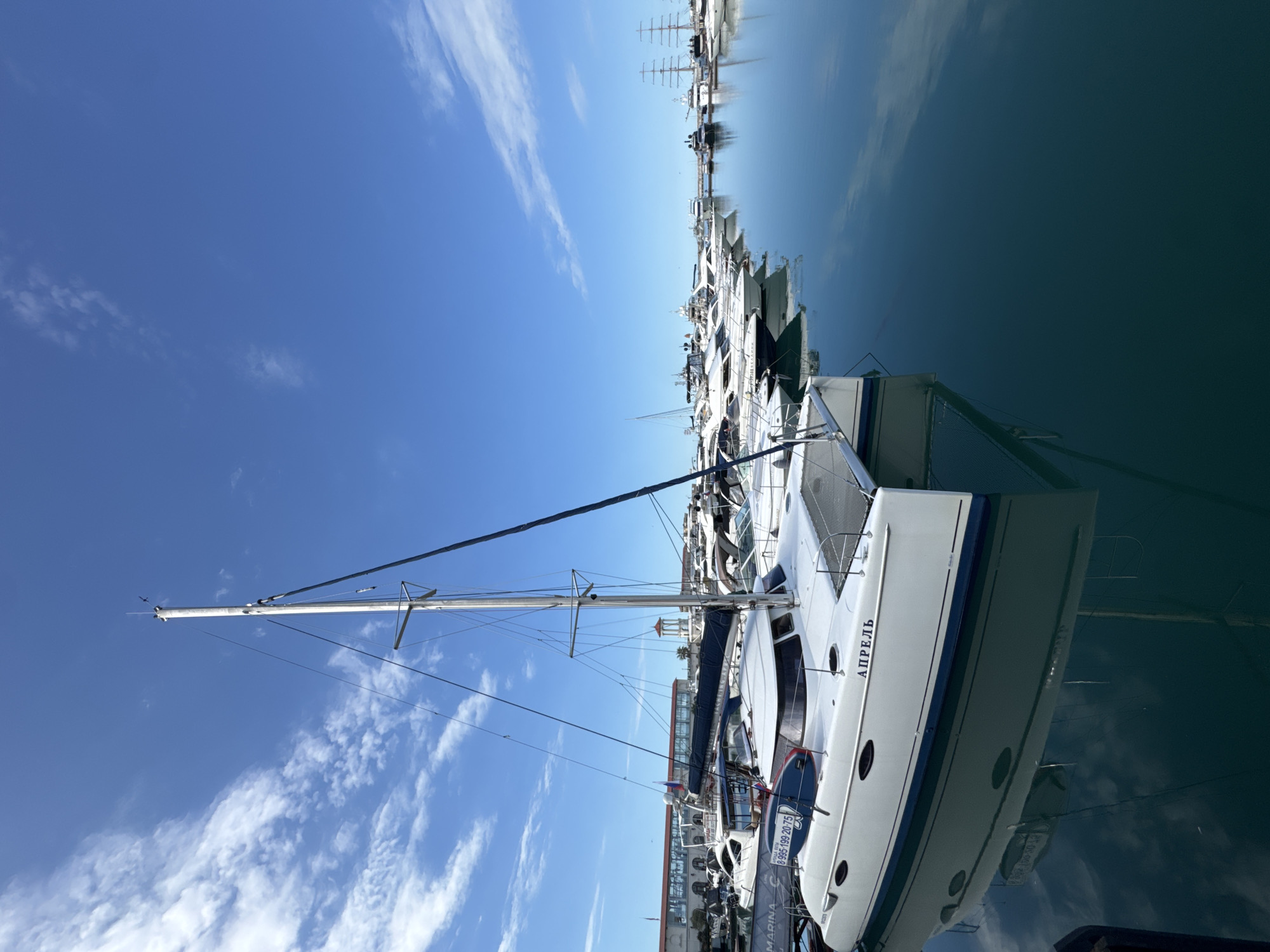 The image depicts a marina in Sochi, Russia, under a clear blue sky with scattered white clouds. The foreground features a white catamaran with the name "АПРЕЛЬ" (April in Russian) visible on its hull. The boat is docked alongside other yachts and vessels of various sizes. The calm, turquoise water reflects the boats and the sky. In the background, several buildings, including structures that appear to be port facilities and possibly hotels, line the waterfront. Tall masts of sailboats and the rigging of larger ships are visible further out in the harbor. The overall scene conveys a sense of a peaceful and sunny day at a bustling marine port.