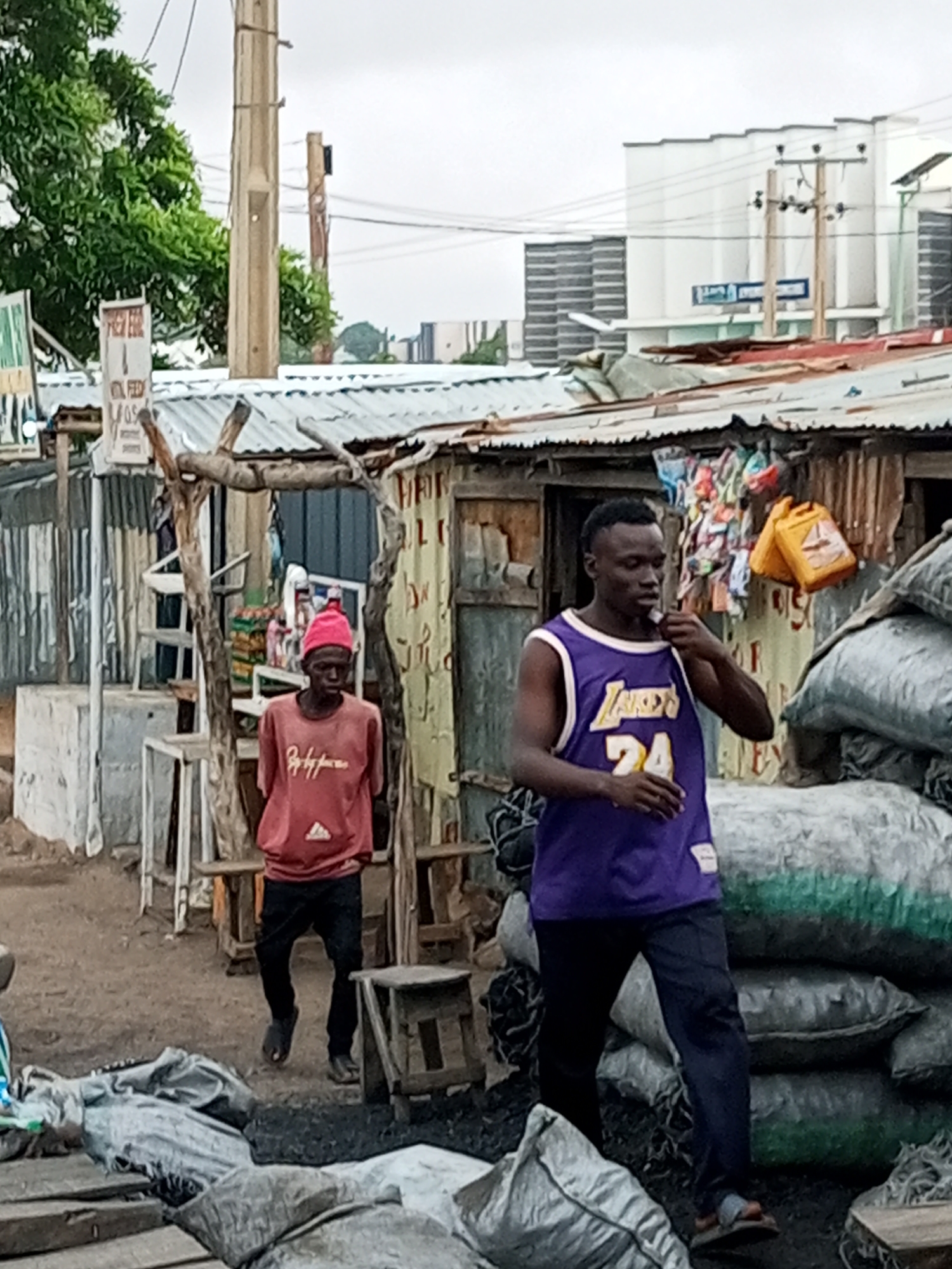 This image depicts an outdoor scene, likely in Tumfure, Nigeria, characterized by its informal setting and a sense of bustling activity. Two individuals are the primary focus. The foreground is dominated by large, dark sacks, presumably filled with charcoal, stacked and scattered across the ground, suggesting a marketplace or a storage area for such goods.
In the middle ground, a young man wearing a purple Los Angeles Lakers basketball jersey with the number 24 is standing and adjusting the collar of his shirt. He has short, dark hair and is wearing dark trousers and flip-flops. To his left, and slightly further back, is another person wearing a red long-sleeved shirt and a pink beanie. This individual is wearing dark trousers and appears to be walking away from the viewer.
Behind the individuals, makeshift structures constructed from corrugated metal and wood are visible, functioning as stalls or shops. These structures are adorned with various items for sale, including brightly colored bags and a yellow plastic container. Signs with text are also present on some of the structures, though the text is largely illegible. The environment appears somewhat dusty or muddy, with a muted color palette dominated by earthy tones, grays, and the greens of distant foliage. Overhead, power lines and poles are evident, suggesting the presence of urban infrastructure. The sky is overcast, contributing to the soft lighting of the scene.