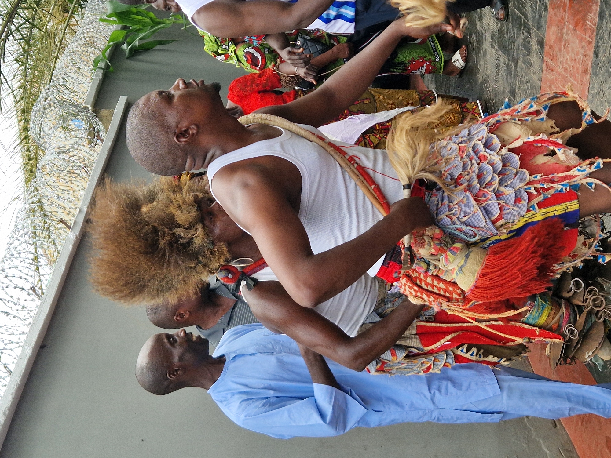 In Kabong, Nigeria, a group of men are gathered outdoors in what appears to be a ceremonial or festive occasion. The central figure, a man adorned in a traditional costume featuring a large, elaborate headdress made of dark, wispy hair, is positioned to the right of the frame. He wears a white tank top and a skirt-like garment made of colorful fabric, adorned with red fringe and intricate patterns. Strapped across his chest is a thick, woven cord. To his left, another man, possibly an elder, stands with his head shaved and a serene expression, wearing a light blue robe with a high collar. Behind him, a third man, also with a shaved head, is partially visible, wearing a gray and white striped polo shirt. In the background, a barbed wire fence runs along the top of a concrete wall, suggesting the event is taking place within a secured or enclosed area. The ground is paved with dark tiles, some of which have red inlays. The overall atmosphere is one of cultural celebration, with the vibrant attire of the central figure being the most striking element of the scene.