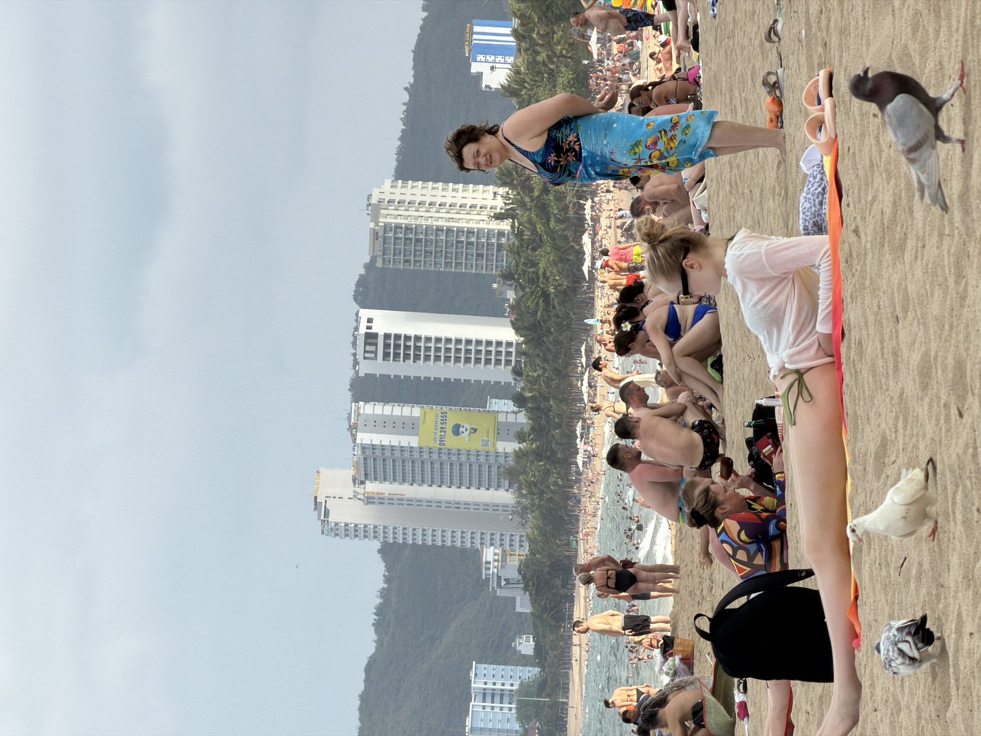 This image depicts a crowded beach scene in Nha Trang Ward, Vietnam. A high density of people are gathered on the sand and in the water. Several high-rise buildings and trees line the background. In the foreground, several people are visible, along with pigeons on the sand. The sky is overcast, suggesting a cloudy day.