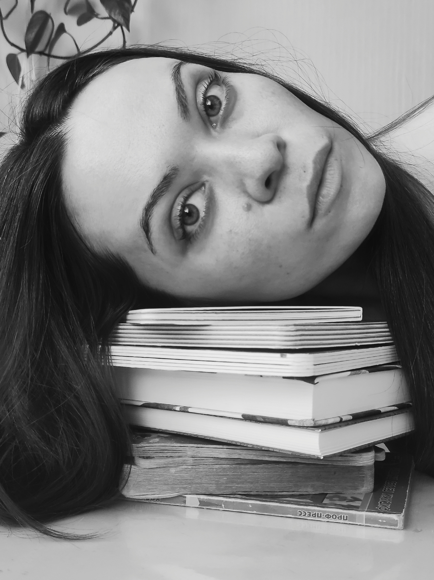 This black and white portrait features a young woman lying on her side, resting her head on a stack of books. Her head is tilted, and her long, dark hair is spread out around her shoulders and the books. She looks directly into the camera with large, expressive eyes. Her face shows a neutral to slightly contemplative expression, with a noticeable beauty mark above her upper lip and a subtle hint of a smile.
The stack of books beneath her head consists of several volumes of varying sizes, suggesting a context of study or quiet reflection. In the background, a houseplant with dark leaves is partially visible, indicating an indoor setting. The lighting creates subtle shadows and highlights, emphasizing the contours of her face and the textures of her hair and the books.
On the spine of the bottom-most book in the stack, Cyrillic text is visible, clearly reading "ПРОФ-ПРЕСС" (PROF-PRESS). Another word or part of a word on the same book, to the right of "ПРОФ-ПРЕСС", appears to be "ФИЗИКА" (PHYSICS). The image captures a moment of quiet introspection or pause, perhaps during a study session. The absence of color adds to the thoughtful and somewhat timeless quality of the scene.