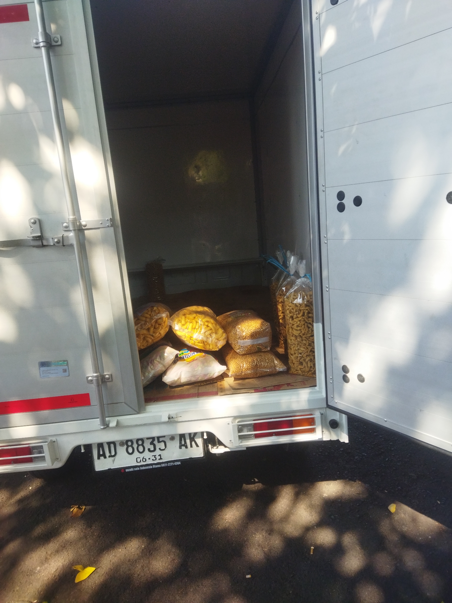 The image shows the open rear of a white cargo truck, parked on an asphalt surface that appears to be a road or parking lot. The truck's interior is filled with several large, clear plastic bags of snacks. Some bags contain a light yellow, irregularly shaped snack, while others contain a macaroni-like snack. The truck has a license plate that reads "AD 8835 AK" and "06-31". A sticker on the back of the truck below the license plate says "Suzuki Solo Indonesia Utama 021-2221-4384". The scene is illuminated by sunlight, casting dappled shadows across the ground and the truck. The overall impression is a delivery truck being loaded or unloaded in an outdoor setting, likely in Wonogiri, Indonesia, given the license plate format.
