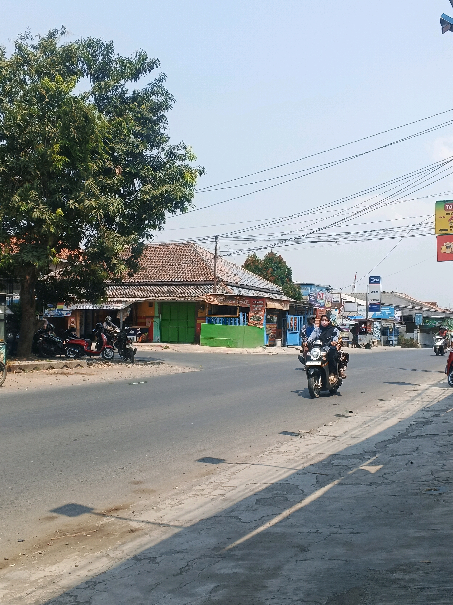 The image depicts a street scene in Taman, Indonesia, under a clear blue sky. A large tree with dense green foliage is on the left side of the frame, casting shadows onto the road. Several motorcycles are parked along the side of the road near a building with a tiled roof and green shutters. Further down the street, people are riding motorcycles. One motorcycle in the foreground has a rider wearing a hijab and a mask. Other buildings and various signs are visible along the street, including an ATM sign. Power lines are strung across the sky. The road appears to be asphalt with some patched areas and debris on the side. The overall impression is of a typical day in a bustling Indonesian town.