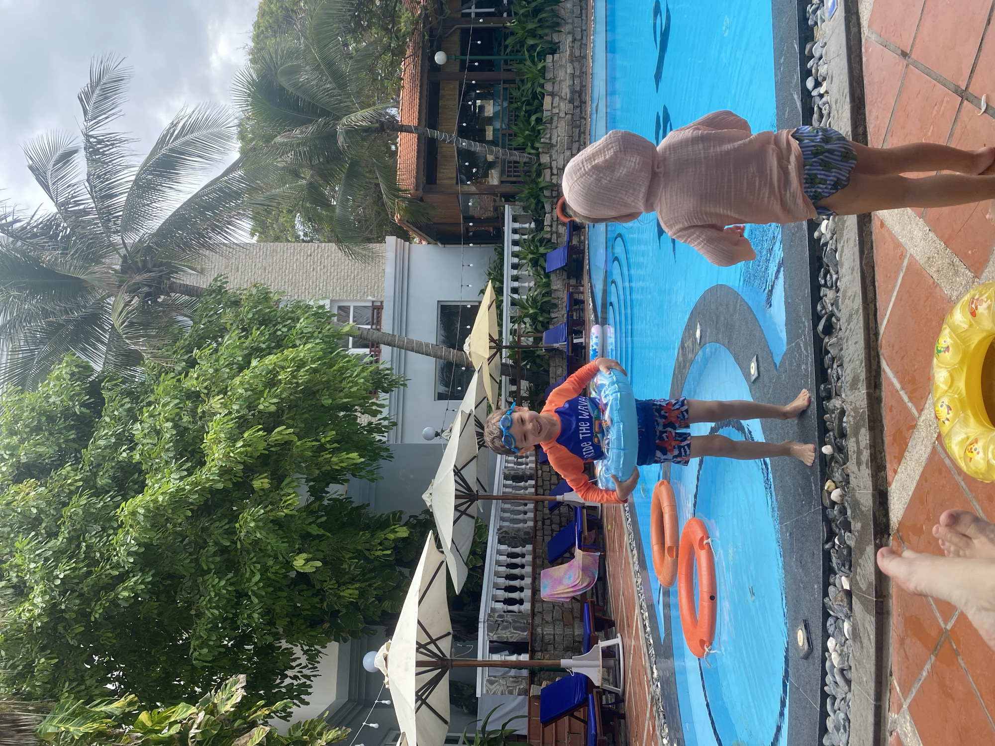 This image depicts a vibrant daytime scene at an outdoor swimming pool, likely within a resort or hotel in Phu Thuy Ward, Vietnam. Two children are visible by the pool. In the foreground, a young boy with brown hair and blue goggles smiles at the camera, holding a blue inflatable ring. He wears an orange and blue long-sleeved rash guard with the text "RIDE THE WAVE" printed on it, paired with patterned swim shorts. Behind him and to the right, another child, viewed from the back, wears a light pink hooded rash guard and patterned swim bottoms, standing at the pool's edge. A pair of adult feet are visible on the terracotta-tiled ground in the bottom right corner, next to a yellow inflatable ring.
The swimming pool features bright blue water with a dark blue patterned design at the bottom and contains two orange lifebuoys. The pool's edge is lined with dark grey tiling and a decorative strip of light-colored pebbles. The surrounding area includes several large, white open umbrellas and blue cushioned lounge chairs, indicating a relaxation space. Lush green foliage, dominated by tall palm trees and dense leafy trees, frames the scene, with resort buildings featuring various architectural styles, including one with a red-tiled roof, visible in the background. The sky is overcast, suggesting a cloudy day. The overall atmosphere conveys leisure and enjoyment at a tropical destination.