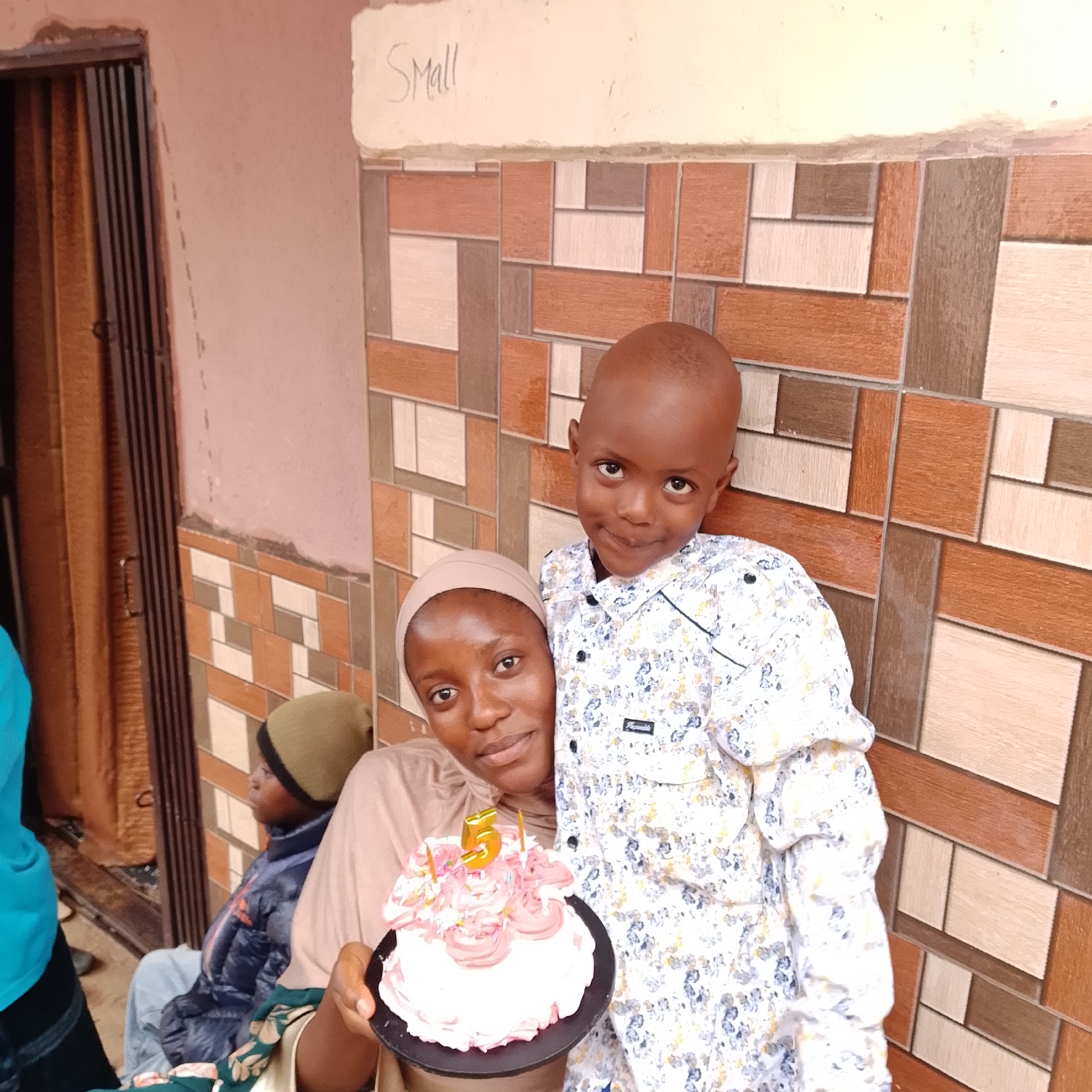 In an outdoor setting with a tiled wall and a closed doorway, a woman and a young boy are posing for a photograph. The woman, wearing a light brown hijab and a tan shirt, is holding a pink frosted cake with a golden number "5" candle and two smaller candles. The boy, wearing a white shirt with a floral pattern, stands behind her. Another person, partially visible in the background, is wearing a dark blue jacket and a green beanie. The wall behind them is adorned with a geometric pattern of rectangular tiles in shades of brown, beige, and white. Above the tiled section of the wall, the word "SMALL" is handwritten in black on a lighter surface. The lighting suggests it is daytime.