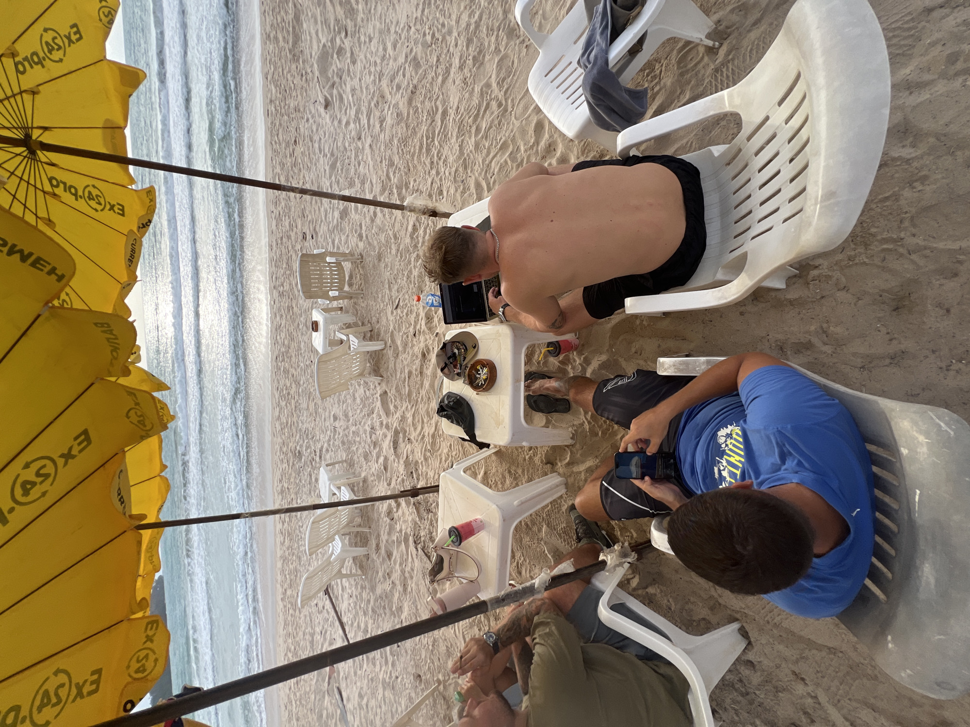 In Choeng Thale, Thailand, on a sandy beach, three individuals are seated under a large yellow umbrella. The umbrella, featuring Thai writing, provides shade for a scene where two men are lounging in white plastic beach chairs. One man, facing away from the camera, is shirtless and wearing black shorts, actively using a laptop on a small white table. Beside him, another man in a blue t-shirt and dark shorts holds a smartphone. A third person, partially visible on the left, is seated in a white chair with a green shirt and grey shorts, their hands positioned as if holding a drink or item. Two small white tables are present, one holding the laptop and the other a dark bag, a hat, and a small bowl with items. The ocean with gentle waves is visible in the background.