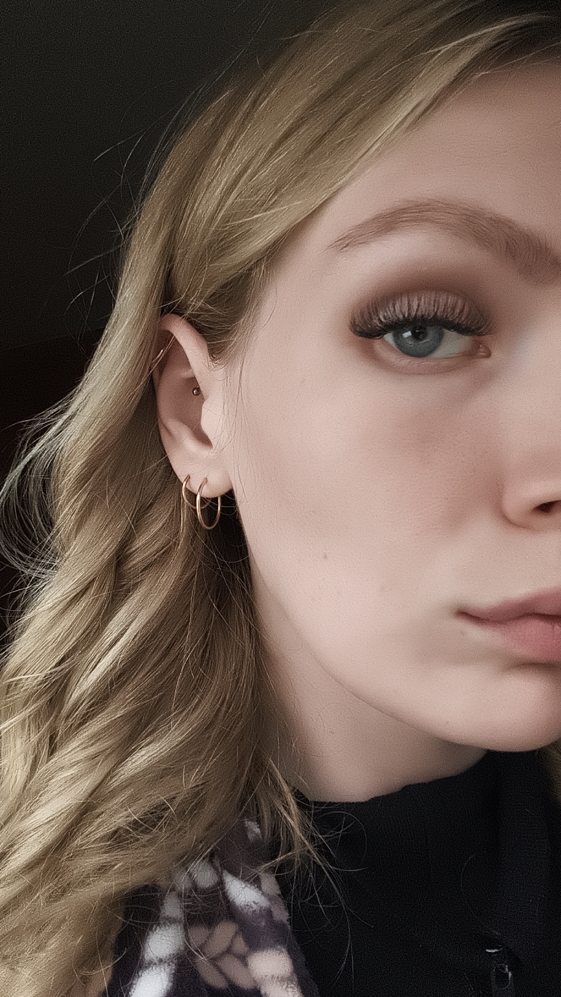 A close-up studio-style portrait captures the left side of a young woman's face. She has light skin, blonde wavy hair, and a light blue eye that is prominently visible with dark, full eyelashes. She has a fair eyebrow and appears to be wearing makeup. Her ear is adorned with three gold hoop earrings and a piercing near the top. She is wearing a black top with a patterned garment underneath. The background is dark. There is no indication of the location being Русский Вожой, Russia, in the photograph.