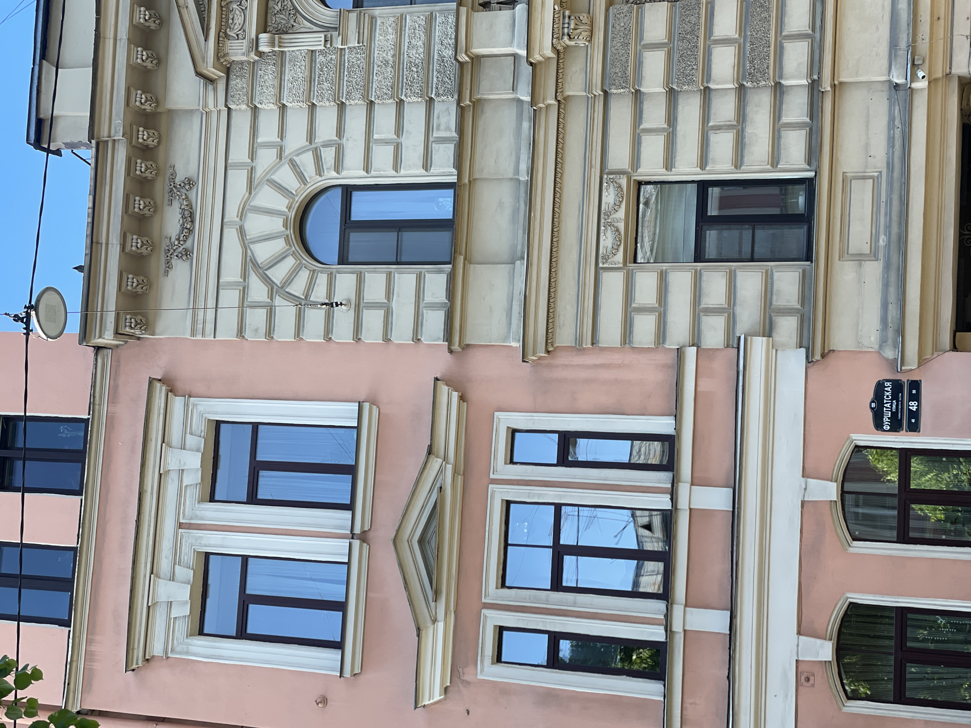 The image shows a building facade in Saint Petersburg, Russia. The building is located on Furstatskaya Street, number 48. The facade is composed of two distinct sections with different architectural styles and colors. The upper section is characterized by light-colored, ornate stonework with rectangular and arched window frames. The lower section features a pink facade with rectangular windows and decorative moldings. The building is several stories tall. The windows reflect the sky and surrounding environment. A street sign is visible on the right side of the facade. A satellite dish is also seen.