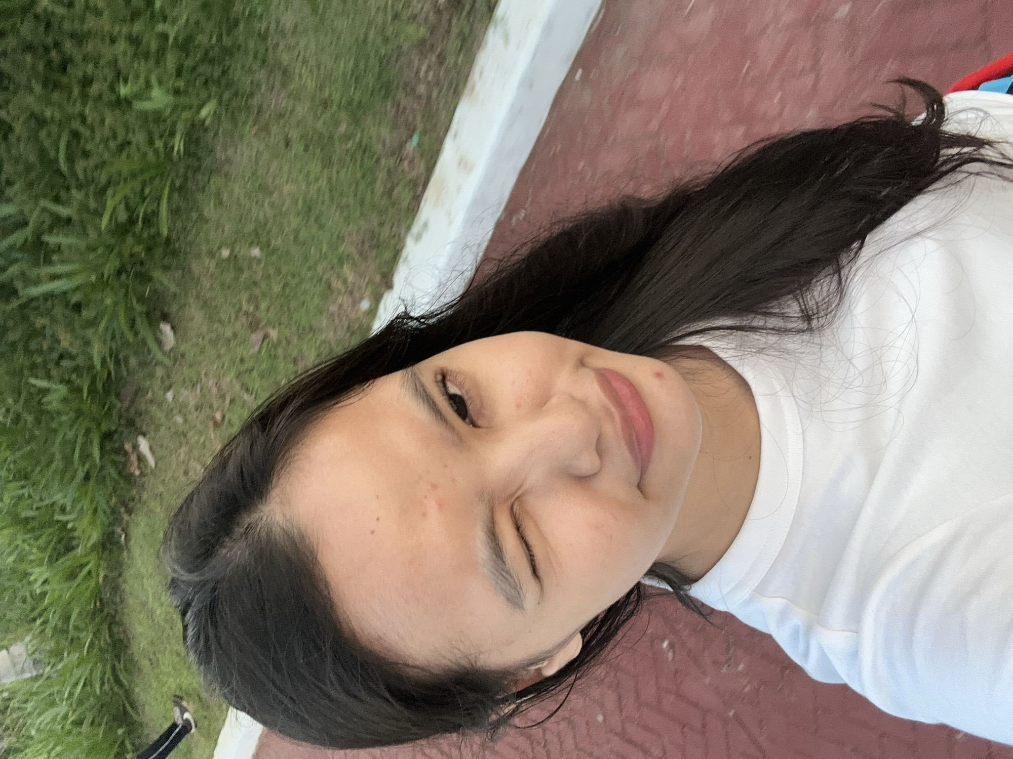 A young woman with dark hair and a fair complexion is taking a selfie outdoors. She is wearing a white t-shirt and winking at the camera. Her face is partially obscured by her hair, which is long and dark. The background shows a grassy area with a paved path and a white border. The scene appears to be a park or a recreational area. This location can be identified as the Davao Recreation Center in Davao City, Philippines, known for its jogging paths and green spaces.