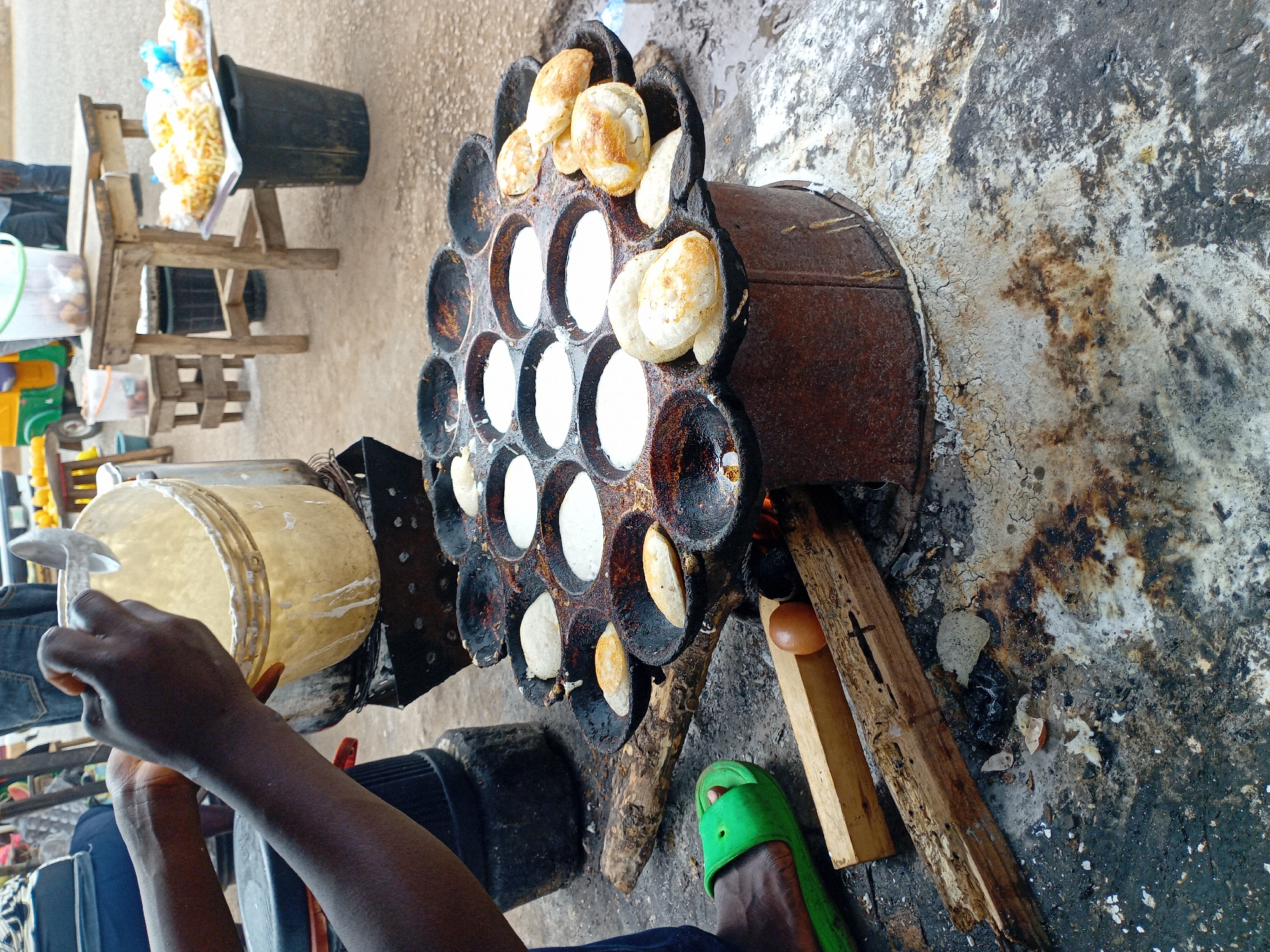 The image depicts a street food vendor in Bukuru, Nigeria, preparing puff-puff, a deep-fried dough snack. A person is shown pouring batter from a translucent bucket with a ladle into a multi-circular cast iron griddle set atop a crude stove, fueled by wood. Some puff-puffs are already cooked, resting around the edges of the pan. A pair of bare feet are seen, one wearing a green flip-flop. To the left is a wooden stool and bucket holding additional packaged items for sale. The ground surrounding the stove is stained, likely from spills and fire residue.