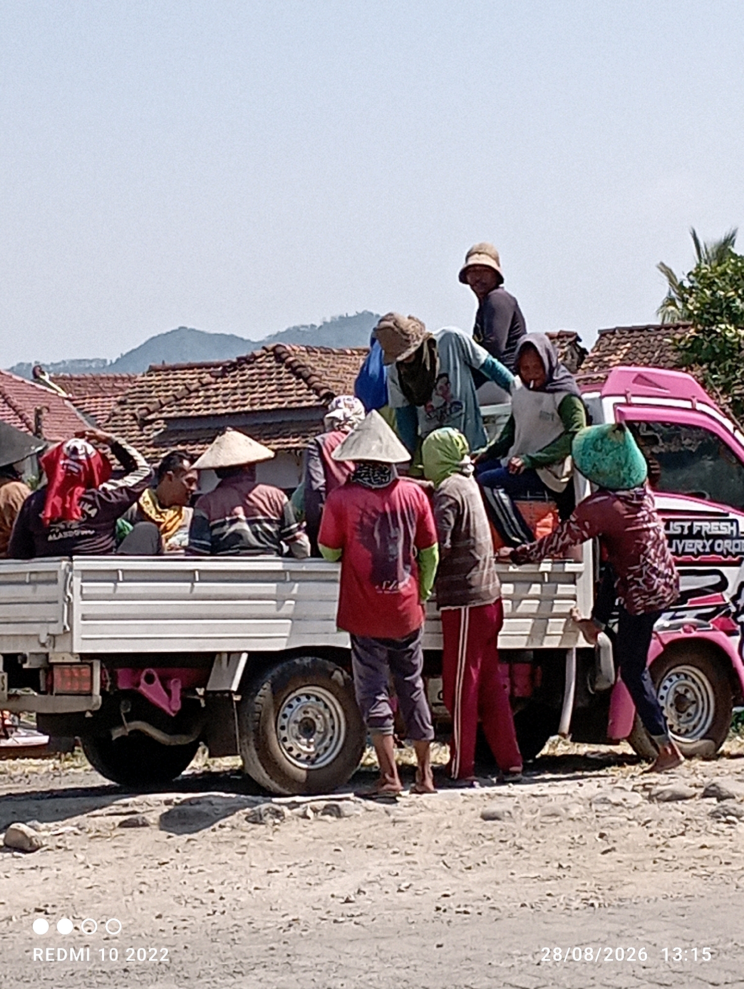 In Karangsari, Indonesia, a group of people are gathered around and on the back of a white pickup truck. The truck is parked on a dirt road, with a pink and white graphic overlay on the side of the truck's cab, featuring text that appears to read "LIST FRESH DELIVERY ORDER". Several individuals, dressed in casual clothing and head coverings, are either standing on the truck bed or disembarking. One person in the foreground wears a red shirt and red pants, while another to their right wears a patterned red long-sleeved shirt and dark pants. A person on the truck bed wears a light blue shirt and dark pants. Another individual with a straw hat is visible in the background. The environment is sunny, with a hazy sky and distant hills and tiled rooftops of houses. The ground is covered with small stones and dirt. The time stamp "28/08/2026 13:15" is visible in the lower right corner.