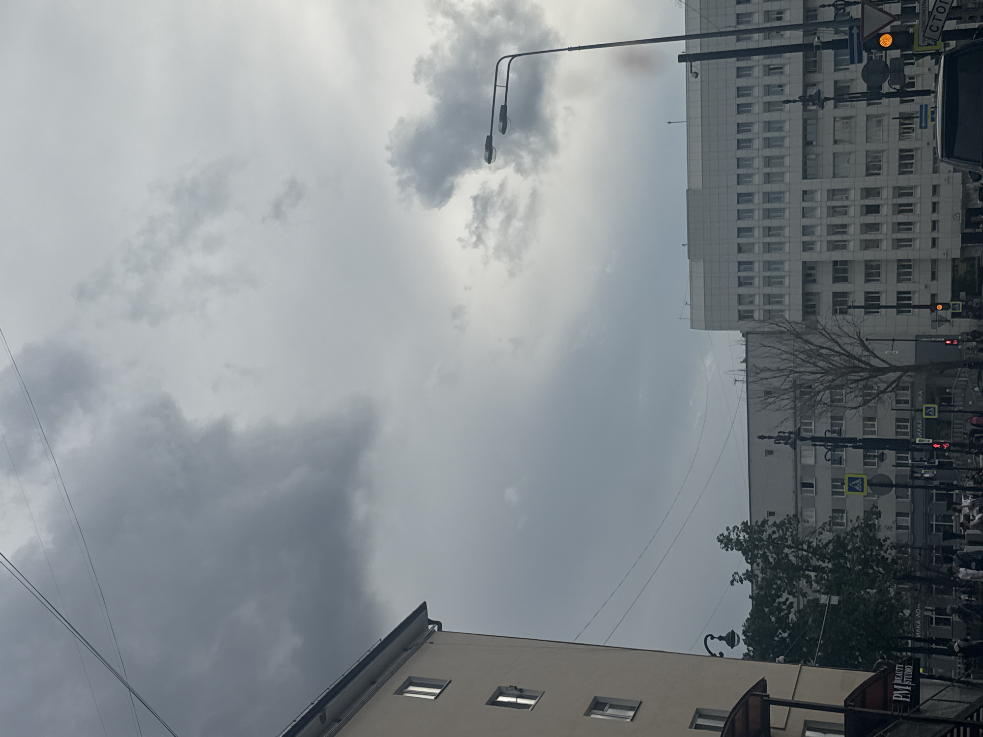 The image depicts a street scene in the city of Perm, Russia, under an overcast sky with dramatic clouds. In the foreground left, a light-colored, multi-story building with several visible windows stands prominently. A sign reading "PM BEAUTY STUDIO" is affixed to the lower part of this building. Adjacent to it, a street lamp with a curved arm is visible.
Further into the scene, a large, white, modern building with numerous windows spans across the mid-ground. Trees with sparse foliage are situated between these buildings, suggesting an autumn or early spring setting. Several streetlights and traffic signals are present, with one signal displaying a yellow light. Pedestrians are visible on the sidewalk and crossing the street. In the distance, more buildings and street infrastructure can be observed. The overall atmosphere is muted, consistent with cloudy weather.