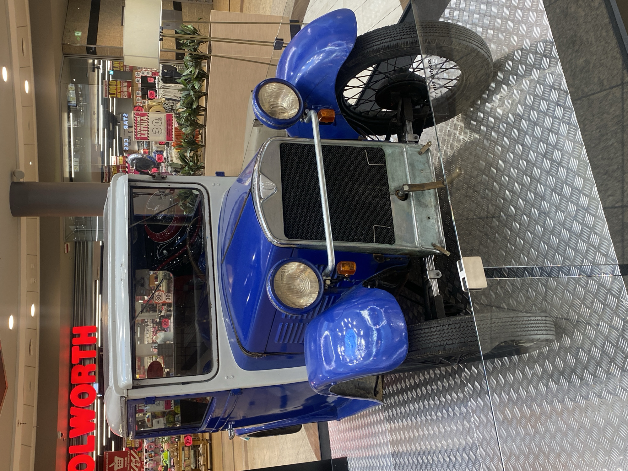 This is a photograph of a vintage blue and white vehicle on display inside a shopping mall. The vehicle appears to be a small truck or delivery van, with a large front grille, two prominent headlights, and a spare tire mounted on the side. It is positioned on a diamond-plate metal platform.
In the background, a shop called "WOOLWORTH" is visible, with signs indicating sales and "LETNIA" (summer) and "BIELIZNA" (lingerie). The overall setting is a bright, indoor retail environment, likely a mall in Łódź, Poland, given the language on the signs. The photograph is taken from a slightly elevated angle, looking down at the vehicle. There are no people visible in the image. The lighting suggests it is daytime, as is typical for a shopping mall. The overall mood is neutral, focusing on the display of the vintage vehicle within a commercial space.