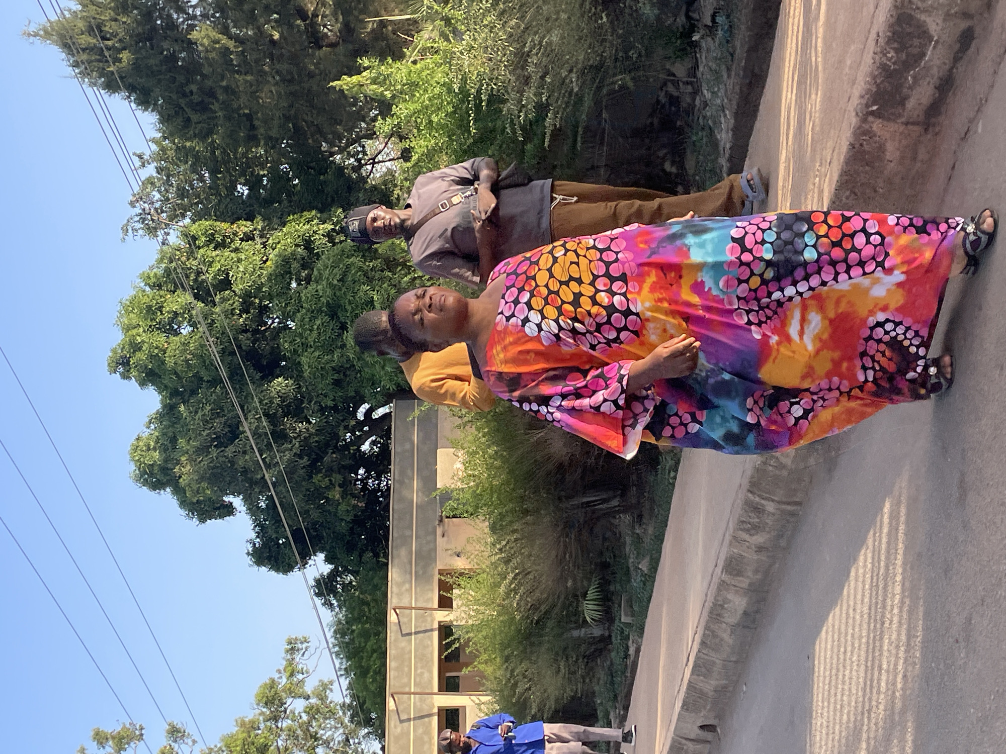 A photograph depicts a group of four people standing on a sidewalk in Jos, Nigeria. The main subject is a woman in the foreground, wearing a colorful, flowing dress with large polka dot and tie-dye patterns. Behind her stands a man in a gray shirt and brown pants. A third person in a mustard-colored shirt is partially visible, positioned between the woman and the man. Further back, a fourth person dressed in a blue outfit is present. The scene takes place on a concrete sidewalk, adjacent to a paved road. In the background, there is a two-story building with simple architectural design. Trees and vegetation are visible, adding a natural element to the urban environment. Overhead, power lines are strung across the clear, blue sky.
