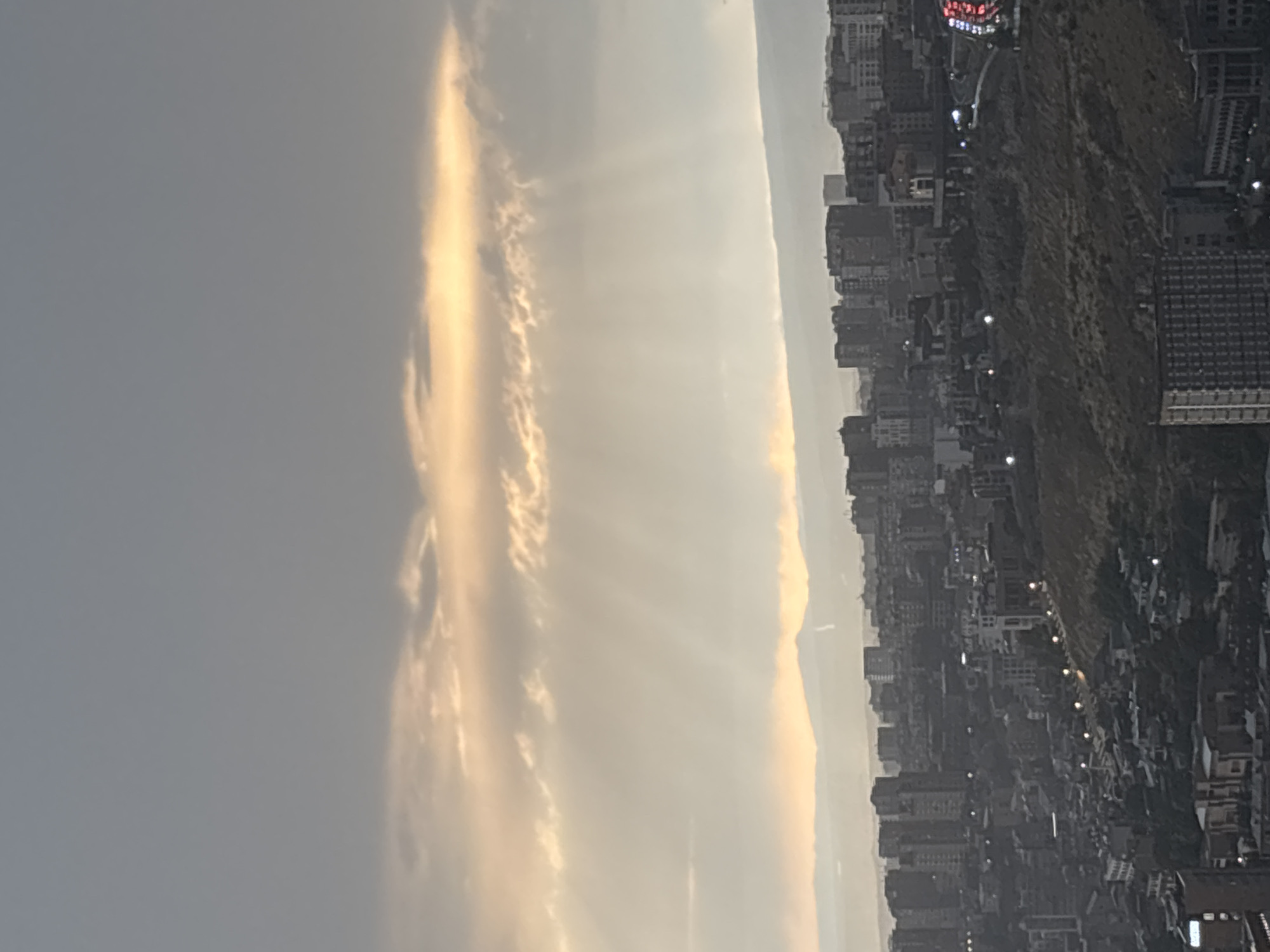 This is a wide shot photograph taken from a high vantage point overlooking the city of Yerevan, Armenia. The sky is overcast with a sliver of golden sunlight breaking through the clouds near the horizon, casting visible sunbeams downwards. In the distance, a hazy mountain range forms the backdrop. The foreground is dominated by a dense urban landscape of buildings of varying heights, ranging from apartment blocks to smaller structures. The city is built on a hilly terrain, with visible roads and streetlights. The overall mood is somber and somewhat muted due to the cloudy sky, but the sunlight offers a touch of hope or drama to the scene.