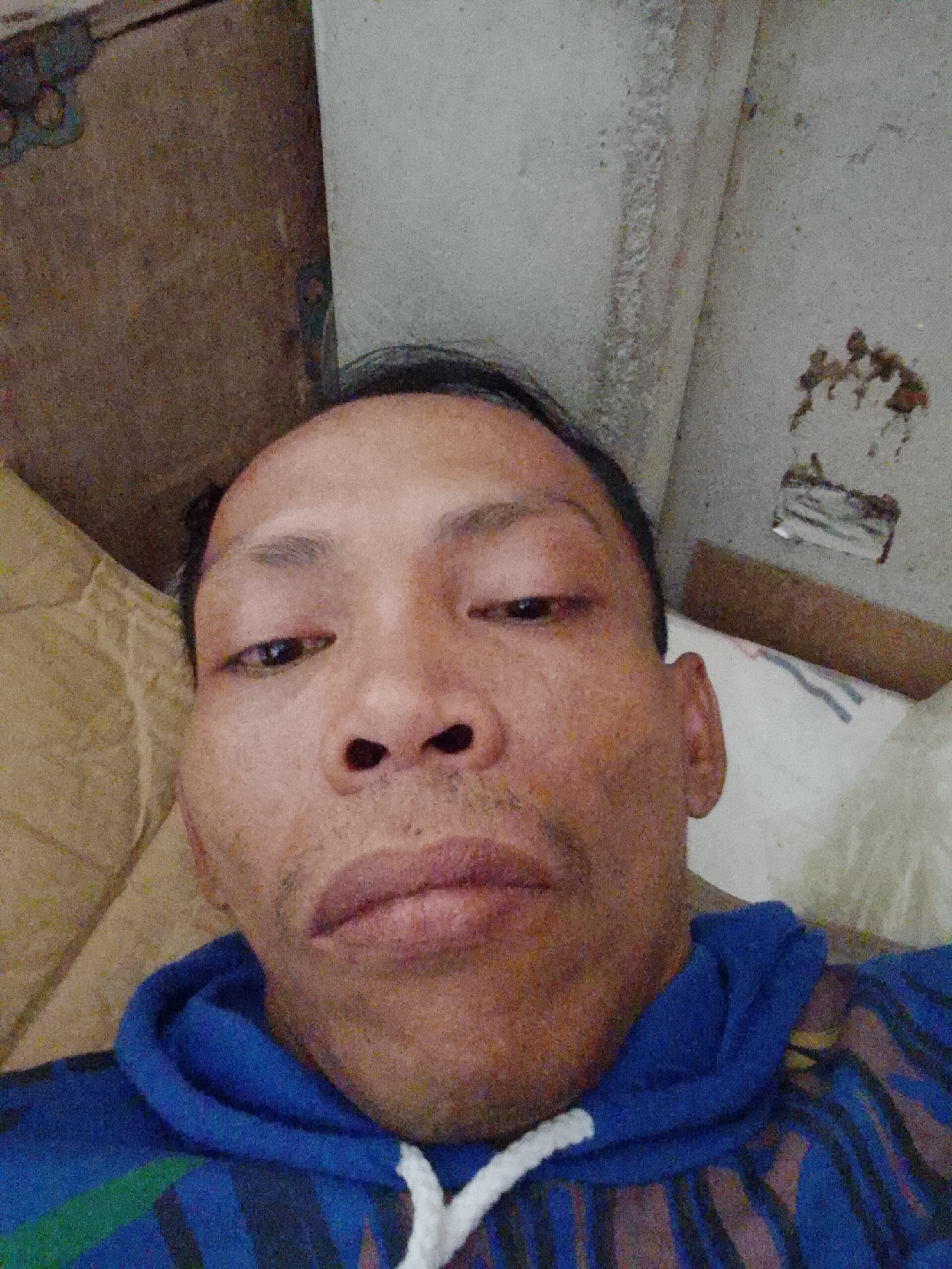 A close-up selfie depicts a man with dark hair, brown eyes, and a slightly furrowed brow. He is wearing a blue and black striped hoodie with a white drawstring. The background is cluttered and includes a beige surface to the left, a textured gray surface above, and a white surface with some debris to the right. There is not enough information to determine the precise location in Bukit Ketok, Indonesia.