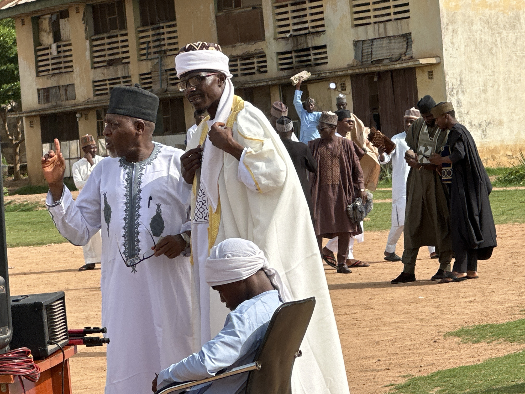 The image shows a group of men gathered outdoors in what appears to be a public space in Kaduna, Nigeria. The men are dressed in traditional Nigerian attire, including robes, caps, and turbans, suggesting a cultural event or gathering. Several men are standing and interacting, while one man is seated in a chair. Behind them is a multi-story building with multiple louvered window panes. A speaker and wires are visible near the foreground, indicating the potential for speech or music during the gathering. The ground is sandy and grassy, with patches of green. Other indistinct figures in traditional clothing can be seen in the background. The scene is captured in daylight, with clear visibility.