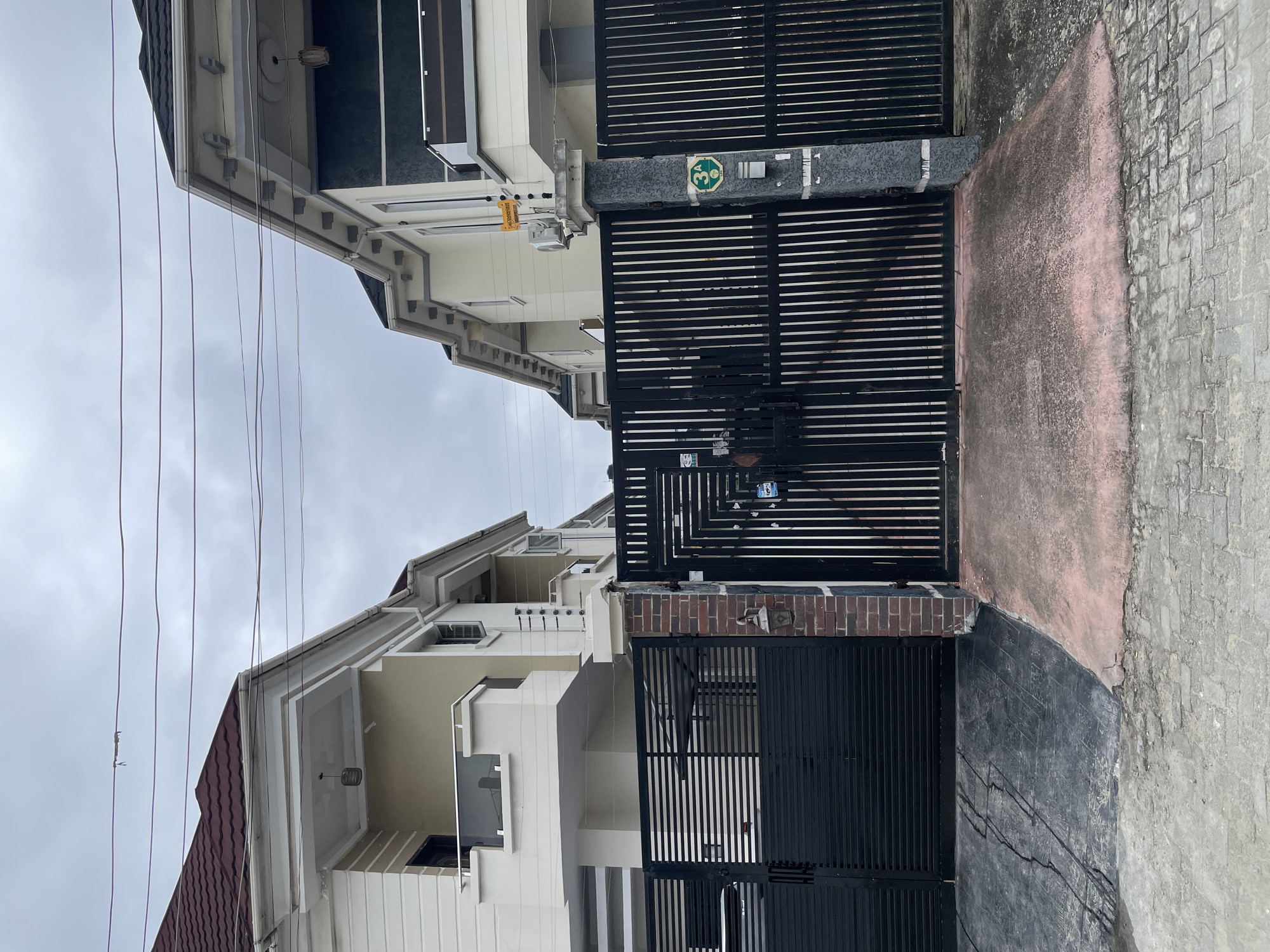 The image depicts the exterior of two adjacent modern residential houses in Ado, Nigeria, under an overcast sky. Both houses are painted in cream and white with contrasting dark roofs. They feature multi-level balconies with glass railings and decorative eaves. The houses are secured by black metal gates. One gate is a solid, horizontal slat design, while the other is a vertical bar design. A brick pillar separates the two gates. On the right side pillar, a green circular sign with the number "3A" is visible, along with a metal mailbox. Electric fence wires are installed on top of the boundary walls of the houses. Power lines are suspended across the sky above the houses and gates. The ground in front of the gates is paved with concrete and stone tiles, showing some signs of wear and tear.