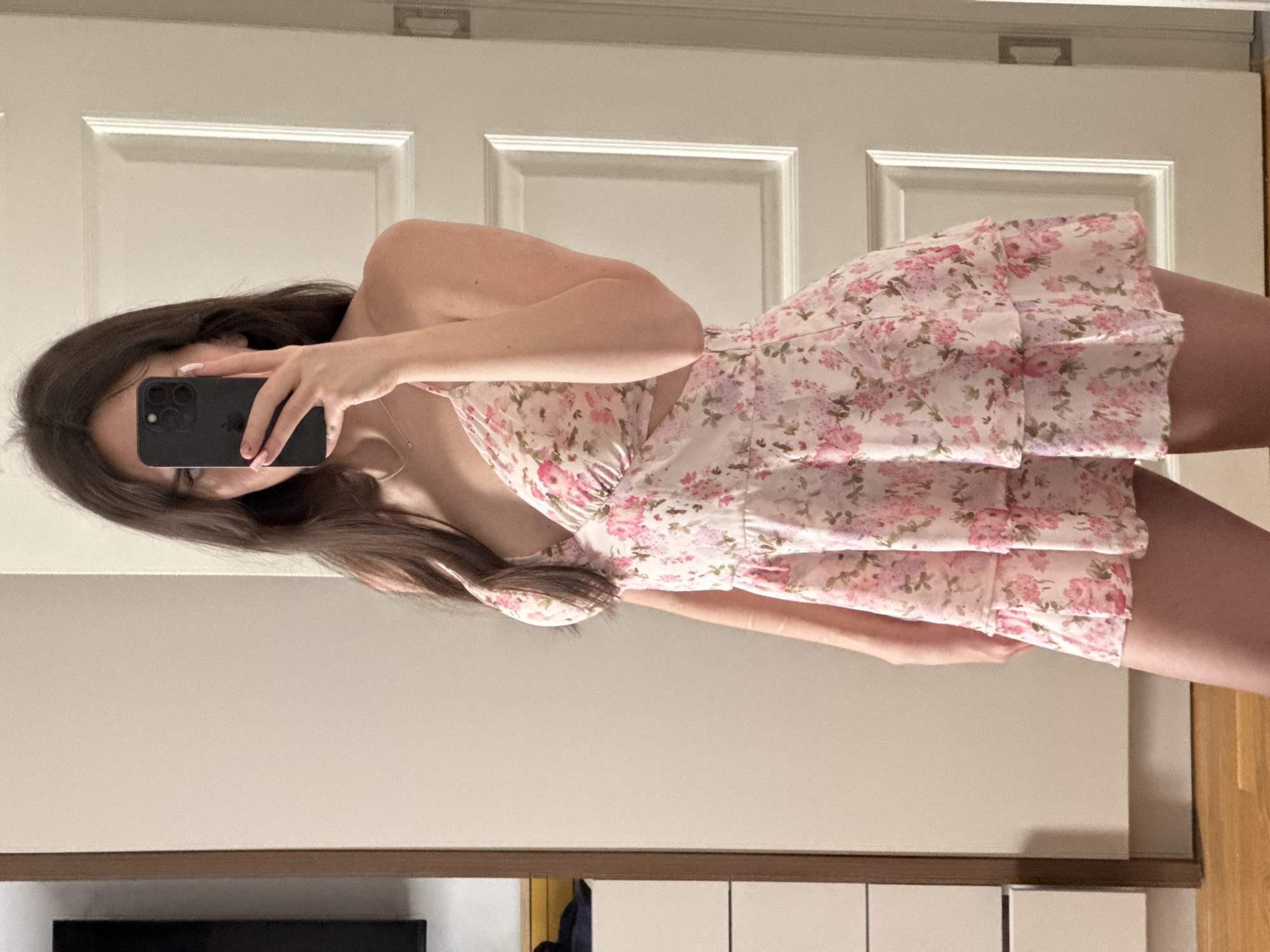 A person is captured in a mirror selfie, positioned sideways in the frame due to the image orientation. They have long, dark brown hair and are wearing a light-colored, possibly white or cream, floral dress or romper. The garment features a dense pattern of pink and purple flowers with green leaves. It appears to be strapless or features thin straps, with a fitted top and a tiered, ruffled skirt section. There's a cutout detail visible on the side of the person's torso.
The person's right arm is raised, holding a black smartphone (likely an iPhone given the three camera lenses) which covers the majority of their face, obscuring facial features. Their left arm is visible, bent at the elbow. A thin necklace hangs around their neck. The person's nails have a light-colored manicure.
The background consists primarily of an interior space. A white paneled door occupies the upper portion of the frame. Below the door, there is a plain, light beige or off-white wall. At the bottom, a dark brown shelf or piece of furniture is visible, beneath which are hints of white panels or drawers and a wooden floor on the far right. The overall lighting suggests an indoor setting, possibly during the day or in a well-lit room. There is no visible text.