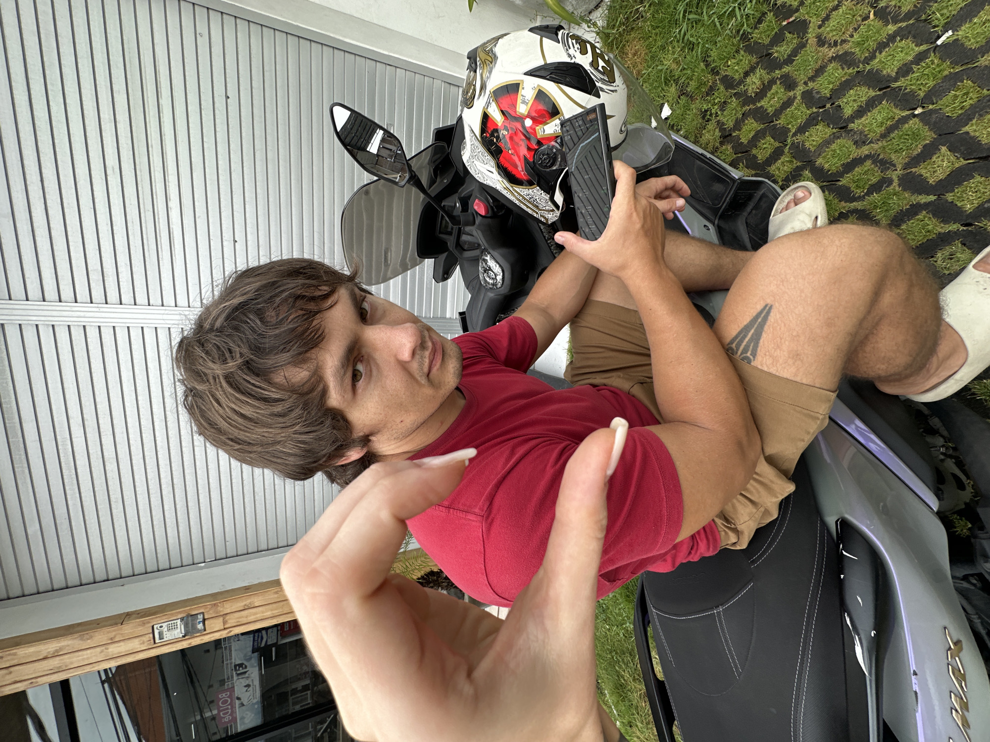 The image shows a man seated on a motorcycle in an outdoor setting. The location is Munggu, Indonesia. The man has brown hair and is wearing a red t-shirt and khaki shorts. He is holding a smartphone in his right hand. A motorcycle helmet is visible on the front of the motorcycle. The background includes a building with a metal roll-up door, and an area covered with grass. A hand with long white nails is in the foreground.