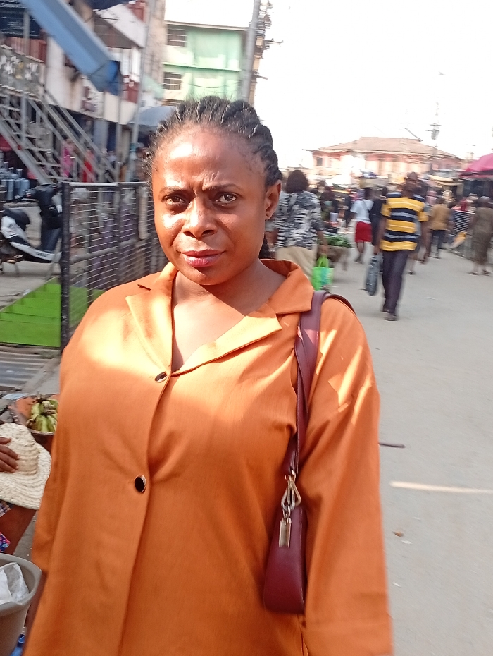 An individual in an orange button-down shirt and a burgundy purse stands in a bustling outdoor market. Buildings and staircases are visible in the background, along with numerous people walking around. The person in the foreground appears to be looking directly at the camera. The atmosphere is bright and seems to be midday.