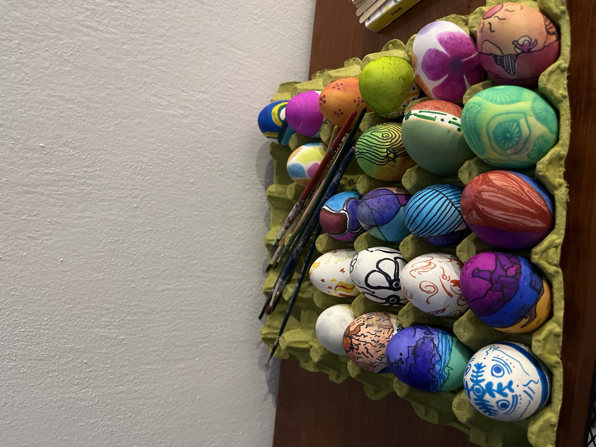 A close-up shot depicts a repurposed green egg carton containing approximately 20 decorated eggs of varying colors and patterns. Paintbrushes are laid across the carton. The carton rests on a dark brown surface, possibly a wooden table or shelf. The background is a plain white wall. The image has an amateur, snapshot quality, with a slightly soft focus. It does not contain any information suggesting a location in San Bernardino Tlaxcalancingo, Mexico.