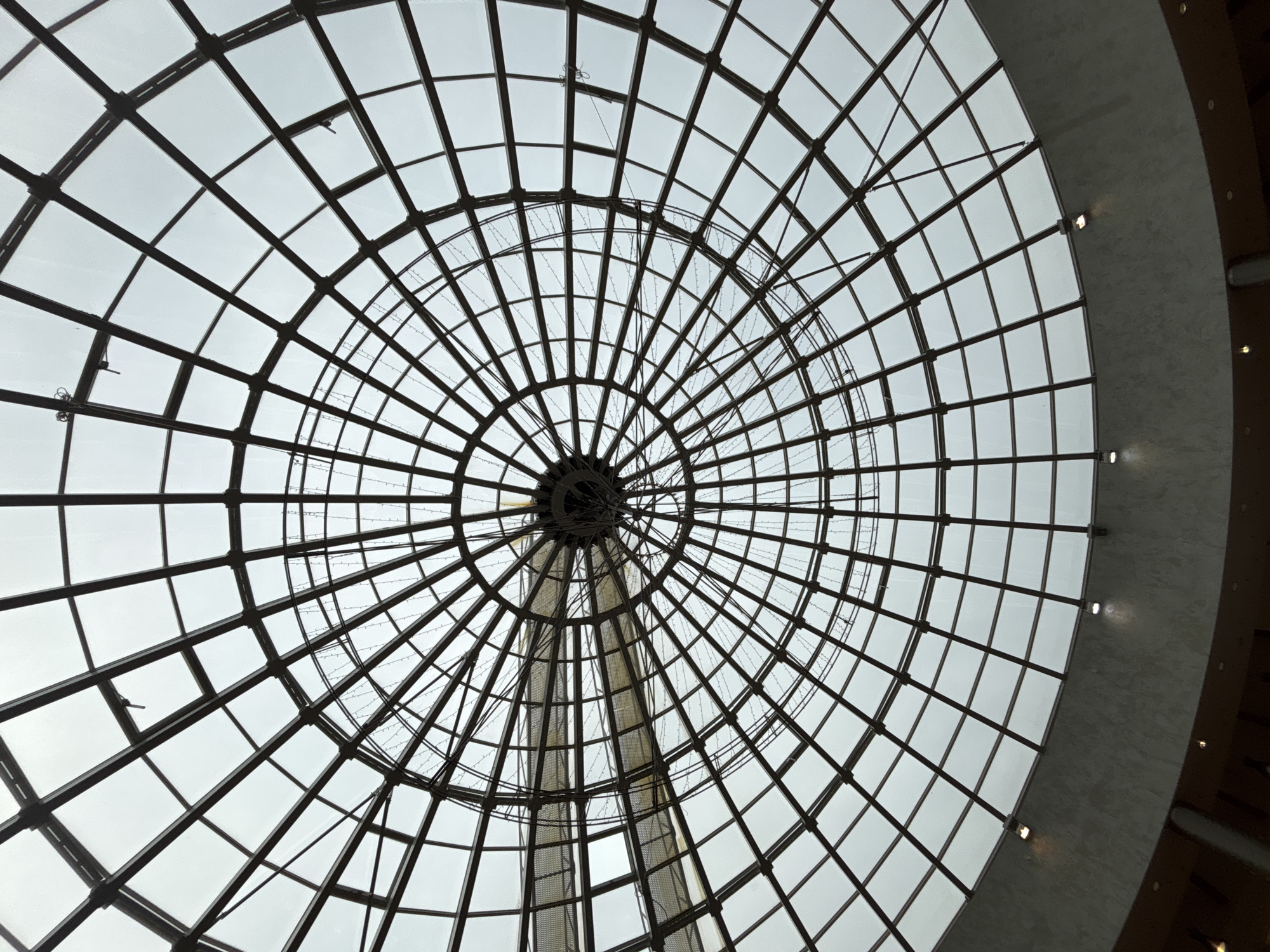 The image depicts an interior view looking directly up into a large, circular, domed glass roof, situated at the Central Department Store (TsUM) in Perm, Russia. The dome comprises a grid of dark-colored structural beams forming radial and concentric patterns, supporting numerous transparent glass panes. Diffused light consistent with an overcast sky illuminates the structure from above. Two curved, elevated walkways or bridges, constructed from a light-colored, possibly perforated metal material, are suspended and traverse the central dome space. Strands of small, decorative lights are draped across the interior surface of the glass dome. The lower perimeter of the dome transitions to a curved, light-grey concrete or plaster wall section, featuring several recessed downlights. Darker architectural elements are visible at the bottom edge of the frame. No individuals are present.