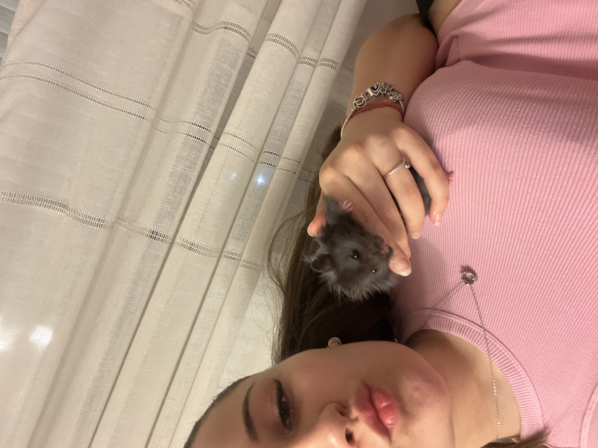 A young woman, with dark hair and light skin, is captured from the chest up in a selfie. She is wearing a pink ribbed tank top and a silver necklace with a pendant. Her lips are pursed, and her left hand holds a small, dark-furred hamster. The hamster is held near her face, and its tiny paws are visible. She is also wearing a silver ring on her left ring finger and a bracelet with charms. The background consists of light-colored curtains, which are partially illuminated by an unseen light source, creating subtle reflections and highlights. The scene appears to be indoors, with soft lighting.