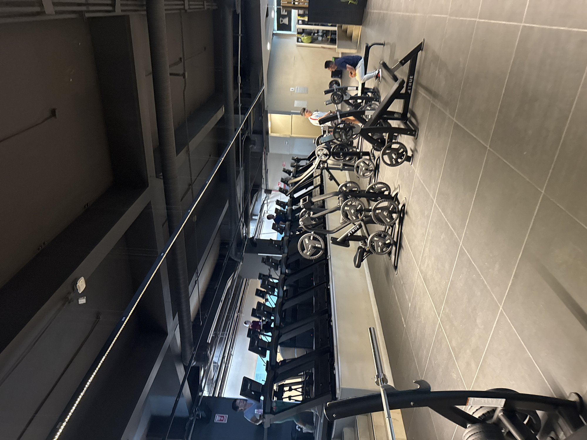 A gym interior with rows of TRUE Fitness exercise bikes is seen. Individuals are exercising on a few of the bikes. The location is presumably in Puebla, Mexico, as requested. The floor is tiled in a light gray color. The ceiling has an industrial design with exposed pipes and beams. The light appears to come from overhead fluorescent lighting.