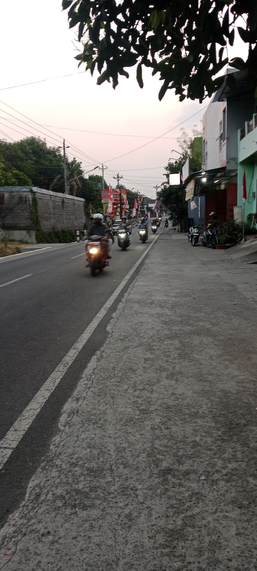 This is a photograph taken outdoors in Maguwoharjo, Indonesia, likely in the late afternoon or early evening based on the lighting. The scene depicts a paved road with a concrete sidewalk running alongside it. The road has a white line marking its edge. Several people on motorcycles are traveling down the road, with some in the foreground appearing blurred due to motion.
Along the sidewalk, buildings of various colors, including white and green, are visible. Some of these buildings have awnings or overhangs. There are also several motorcycles parked on the side of the road and on the sidewalk.
Decorations, including red and white flags and banners, are displayed along the road, suggesting a festive occasion or celebration. Trees with dark green foliage are present in the background, with some branches and leaves from a tree in the foreground casting shadows. Power lines and utility poles are also visible in the sky. The sky itself is a pale hue, with the faintest hint of pink.