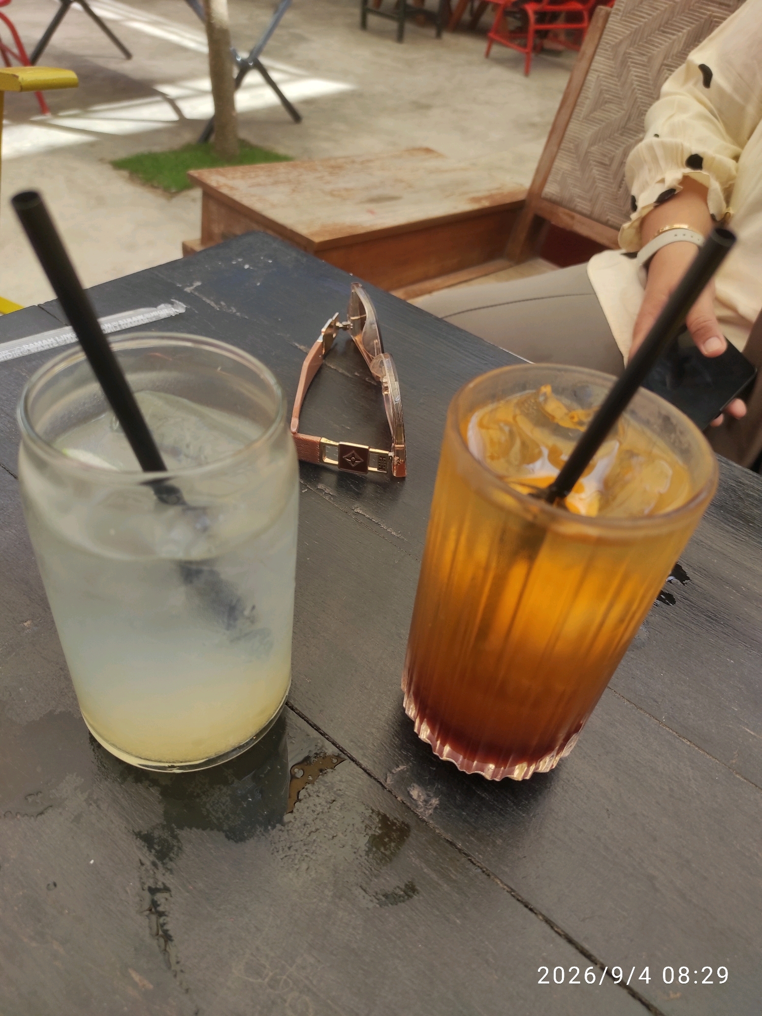 A close-up, eye-level shot shows two iced drinks on a dark, wet wooden table. The drink on the left is in a clear glass with a black straw, appearing light yellow or white. The drink on the right is in a fluted amber glass with a black straw, and is a dark orange color fading to red at the bottom, with visible ice. A pair of rose gold-colored glasses with a brown strap is placed on the table between the drinks. In the background, out of focus, a person's arm wearing a cream-colored blouse with black polka dots is visible, holding a dark smartphone. Further in the background, there is outdoor seating with chairs and tables, suggesting a cafe or restaurant environment. The timestamp "2026/9/4 08:29" is visible in the bottom right corner of the image. The scene is likely a cafe in Trenggalek, Indonesia.