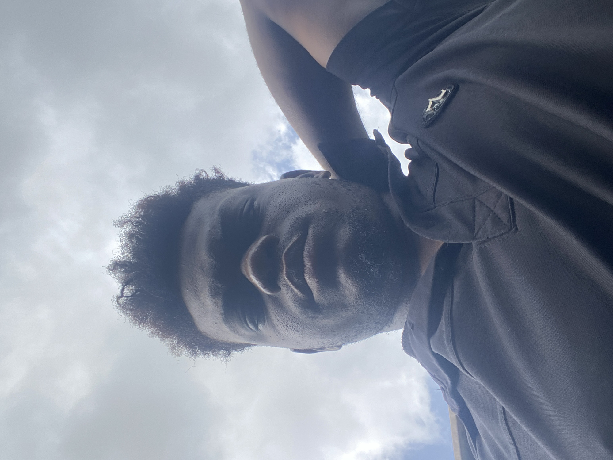 A low-angle shot captures a person with curly dark hair, positioned against a backdrop of a cloudy sky. The individual is wearing a dark polo shirt featuring a small emblem on the upper right side. The shirt's texture and seams are visible. The person appears to be outdoors during the day.