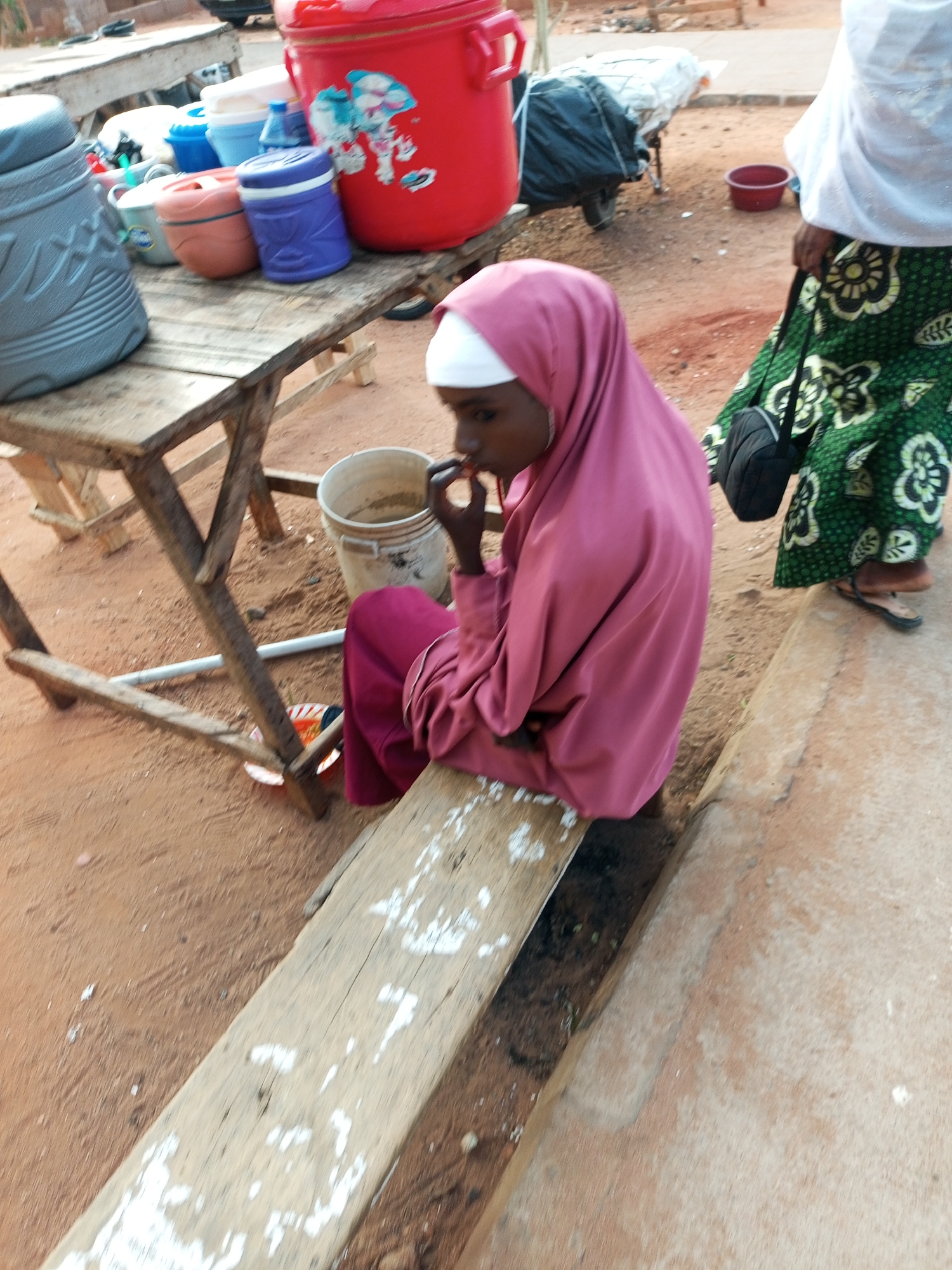 A person wearing a pink hijab and maroon clothing is sitting on a wooden bench, eating. The person is looking down and to the side. A person wearing a green and black patterned skirt and a white top is standing behind the person on the bench. They are standing on a dirt surface. In the background, there are several large plastic containers, including a red one with a blue and white sticker. There is also a motorcycle covered with a white tarp. The image appears to have been taken in a market or outdoor setting, likely in Gombe, Nigeria, given the context. The ground is reddish-brown, consistent with the soil in the region. There are also wooden tables and some buckets visible. The lighting suggests it is daytime, possibly late morning or afternoon.