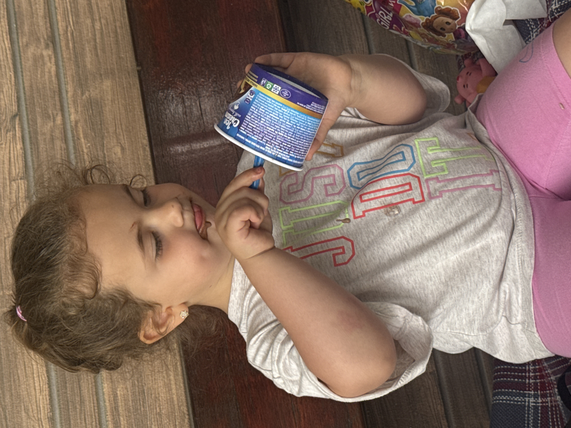 A young Caucasian girl with curly brown hair pulled into a ponytail is seated, holding a small cup of what appears to be ice cream and a blue spoon. She is wearing a light grey t-shirt with colorful lettering that reads "JUST DO IT" and bright pink shorts. Her tongue is slightly sticking out as she appears to be eating the ice cream. She has a small stud earring in her left ear. The background consists of a wooden bench. To the right of the girl, a colorful bag with cartoon characters is partially visible, along with a white tissue and a small pink toy. The lighting suggests an outdoor setting, possibly on a warm day. The image was taken in Rusokastro, Bulgaria.