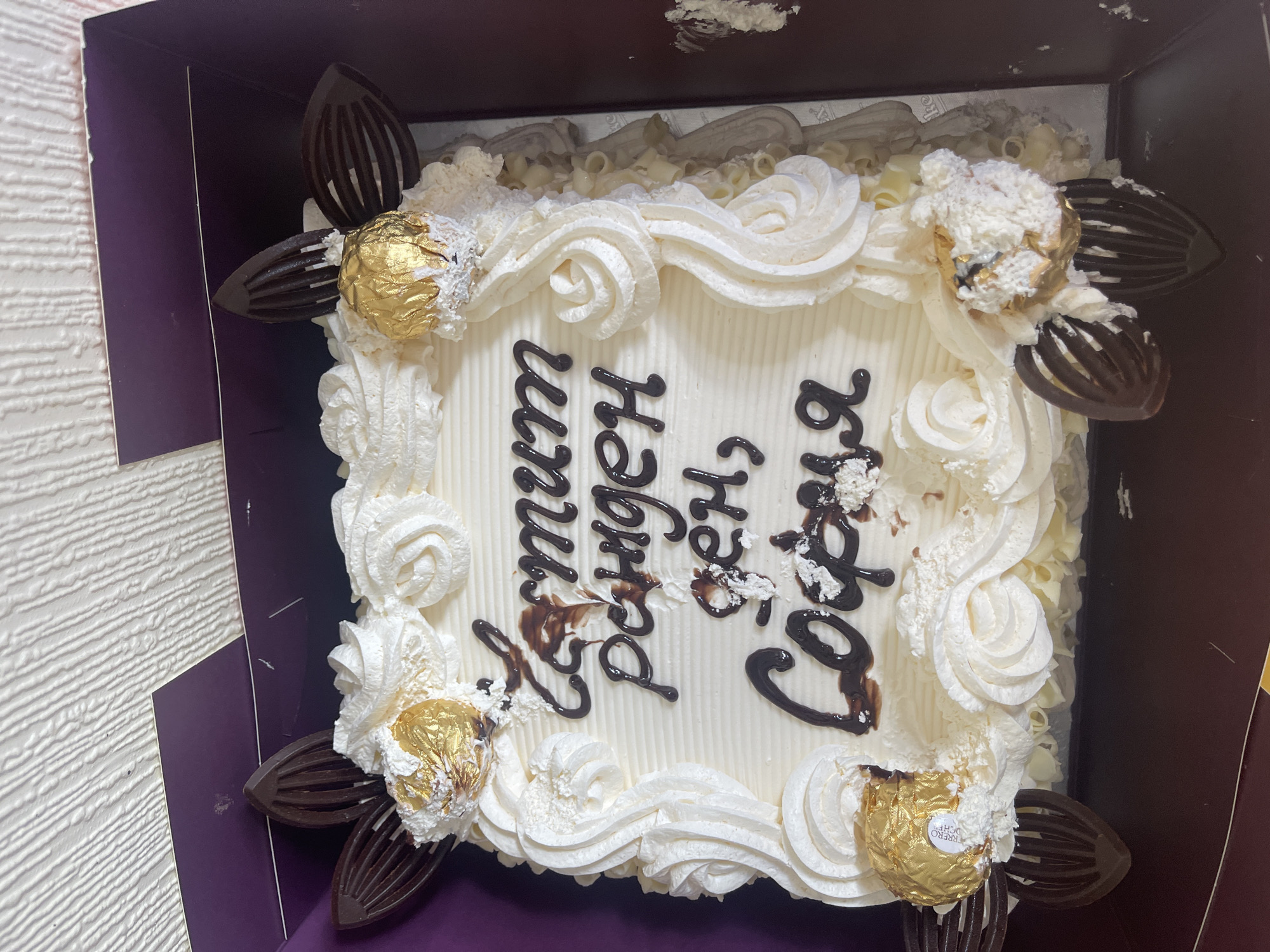 A square cake in a purple cardboard box is positioned in Maidstone, United Kingdom, likely indoors on a table. The cake is adorned with white frosting swirls, chocolate decorations, and Ferrero Rocher chocolates placed at each corner. Dark brown lettering is inscribed on the top surface of the cake, oriented vertically and reading "С Днем Рожденья, София" (Happy Birthday, Sofia). The interior of the box appears dark brown, contrasting with the white frosting of the cake. The background is a white wooden panel, possibly a wall or tabletop.
