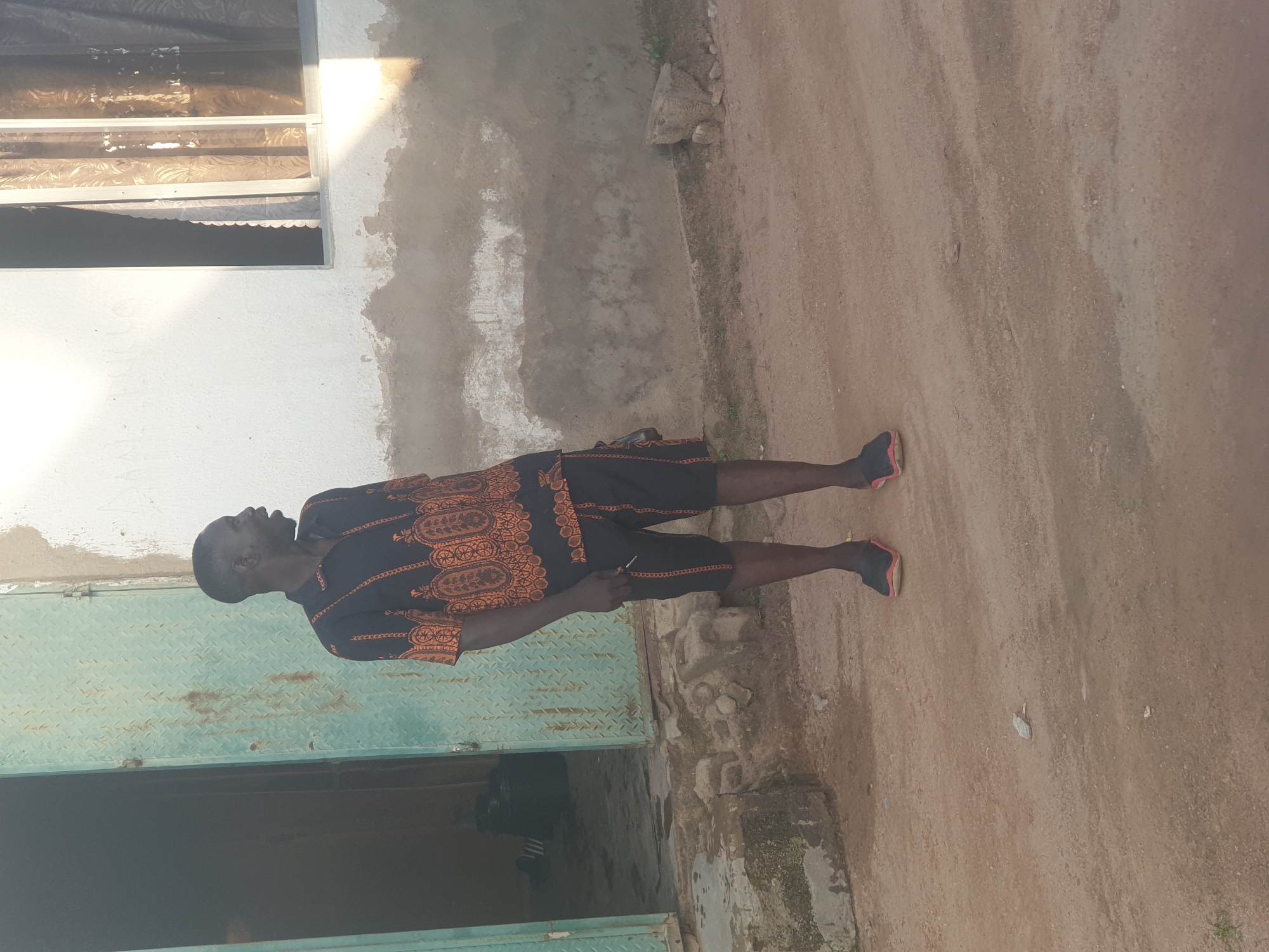 The image depicts a male standing outdoors on a dirt path next to a building in Unknown, Unknown. The individual, an adult male with dark skin and short dark hair, is positioned slightly right of center, facing towards the right with his mouth open and jaw slightly dropped, as if speaking or reacting. He is wearing a black short-sleeved collared shirt and matching black shorts, both featuring an intricate orange-brown geometric pattern. His feet are clad in dark slip-on shoes with a red/pink trim. In his partially visible right hand, he appears to be holding a small, thin, light-colored object, possibly a cigarette or stick.
To the left of the man is a partially open, tall, hinged metal door, painted a light blue-green and exhibiting a diamond-plate texture with some rust. The interior visible through the door is dark. The ground near the door is uneven, wet, and appears muddy with debris. To the right and behind the man is a building with white-painted walls. The lower section of the wall reveals exposed, rough concrete or plaster that is discolored and patchy. A window with a light-colored frame is present on the right side of the building, featuring patterned brown/gold curtains drawn inside. The ground in the foreground and middle distance is a reddish-brown dirt or gravel path, with scattered small stones and debris. Strong, directional sunlight casts prominent shadows on the building's wall and illuminates the scene from the right.