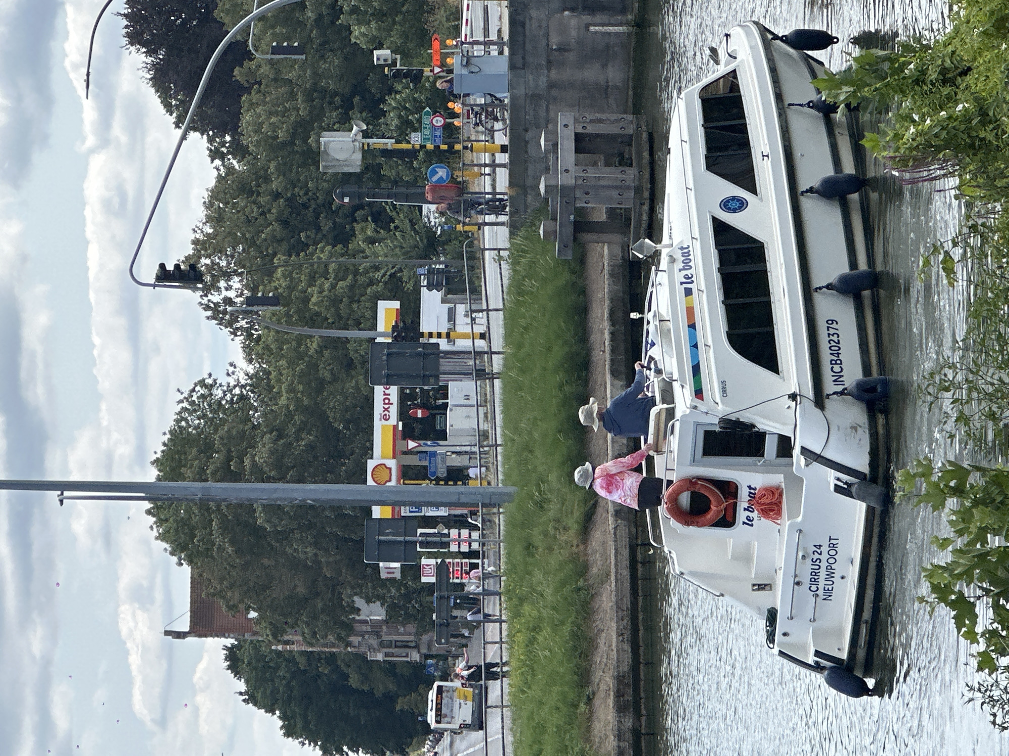 This is a vertical, eye-level, medium shot of a white canal boat docked in a canal with green grassy banks. The boat is the primary subject, with three people visible on its deck, preparing to depart or having just arrived. Two individuals, wearing hats, are on the back deck. One person, wearing a pink and white floral shirt, appears to be adjusting a life preserver. The other person, wearing a blue jacket, is out of focus, but their head and shoulders are visible. The boat has tinted windows and "le boat" is written on its side. The registration number "INC B402379" is also visible on the stern.
In the background, across the canal, is a canal lock with various signal lights and signage. A Shell gas station is also visible, with multiple pumps and signs indicating "express." Traffic lights are visible on the left side of the image, suggesting the proximity of a road. The overall scene appears to be a sunny day with scattered clouds, though the lighting suggests it might be late afternoon or early evening. The presence of the canal lock and the boat with "le boat" branding indicate a location in a region popular for canal tourism, such as Bruges, Belgium. The mood appears relaxed and casual.