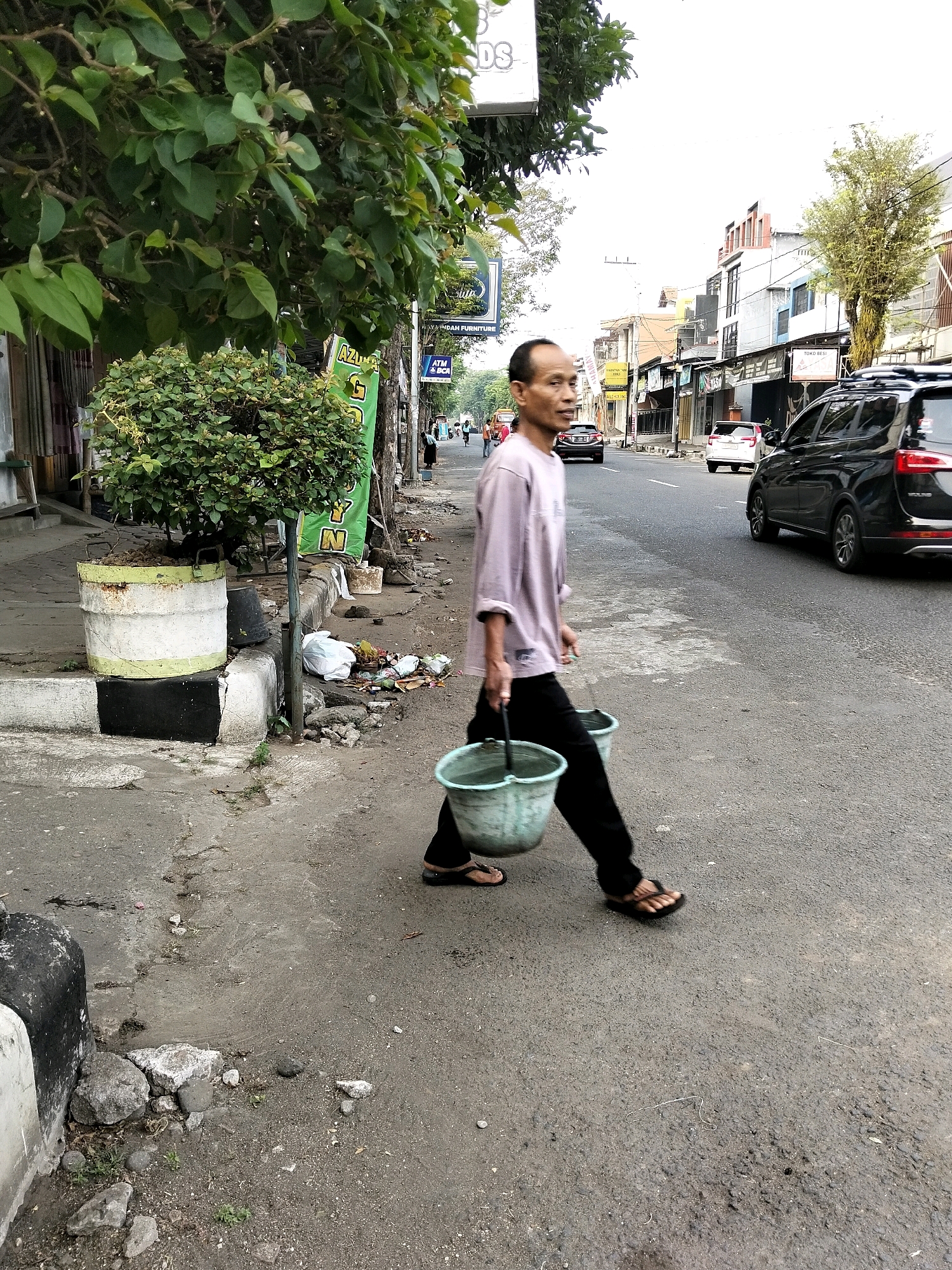 A man is walking on the side of a road in Purworejo, Indonesia, carrying two buckets by their handles. He is wearing a light purple long-sleeved shirt and black pants, and appears to be wearing flip-flops. The buckets are light green. The man is walking on a paved area next to a street with cars driving on it. To his left is a sidewalk with some greenery and a large planter. There are also piles of trash on the side of the road. The background shows buildings and trees.