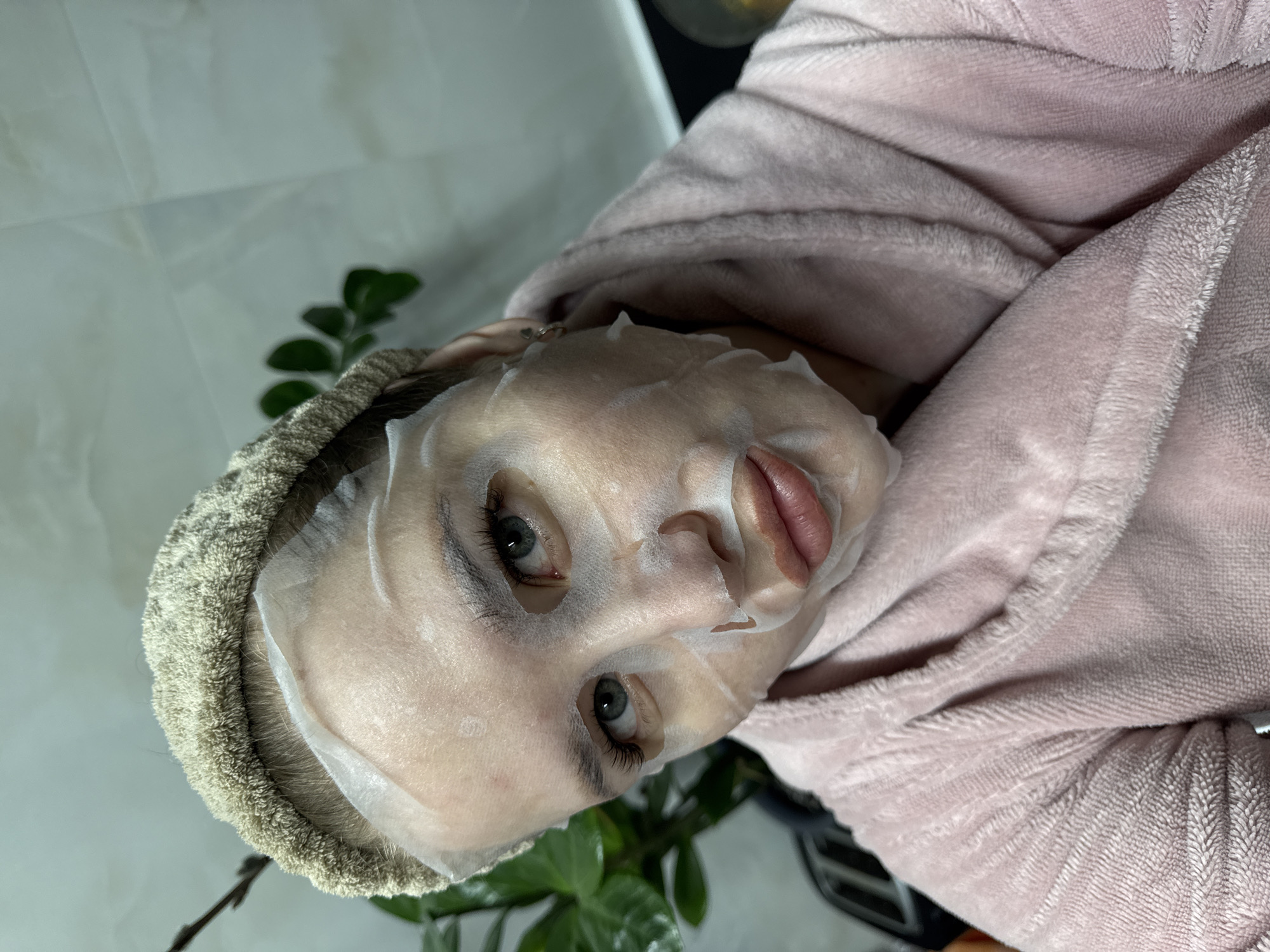 A close-up, eye-level portrait photograph captures a person engaged in a self-care routine, with the image rotated 90 degrees clockwise. The subject, likely female, has a white sheet face mask applied, covering most of her face except for her light blue or green eyes and slightly parted lips. Her eyes have prominent dark eyelashes, suggesting mascara or lash extensions.
She is wearing a soft, fuzzy, light pink or peach-colored bathrobe, which drapes over her shoulders and upper body. A light brown or beige terry cloth headband or towel is wrapped around her head, securing her dark hair.
The background is softly blurred. In the lower left corner (when viewed upright), dark green plant leaves are visible. The rest of the background appears to be a light-colored, indistinct surface, possibly a wall or fabric. The overall scene conveys a sense of calm and relaxation, characteristic of a beauty or wellness activity. There is no visible text in the image.