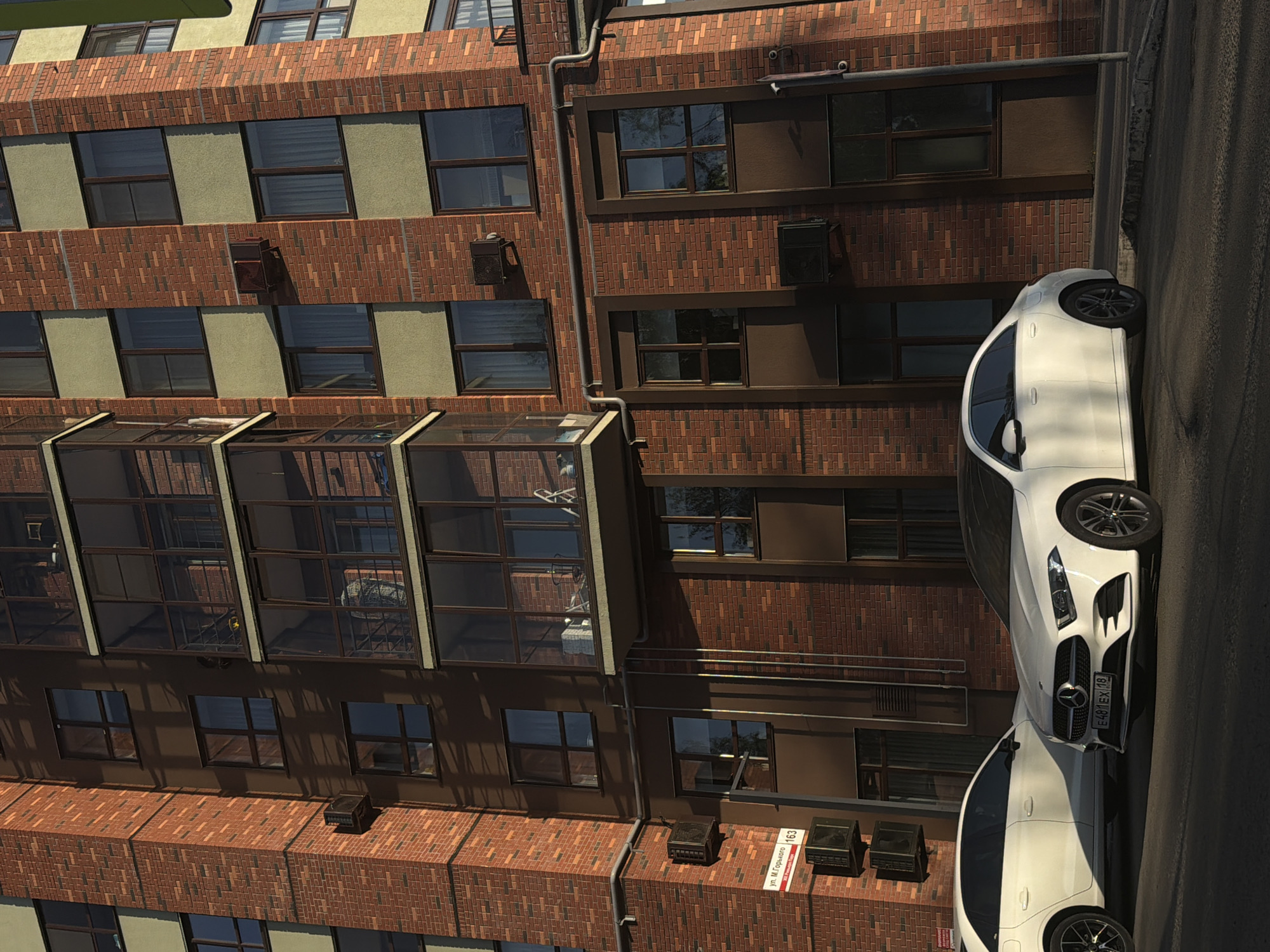 The image captures a scene in Izhevsk, Russia, featuring a view of a red brick building and two white cars parked along the side of the road. The building has multiple stories, with a mix of windows and glass-covered balconies. Air conditioning units are mounted on the exterior walls. A sign with Cyrillic text indicating "ул. М. Горького 163" (Ulitsa M. Gorkogo 163), is also visible, denoting the address as 163 Gorky Street. Two white cars, partially visible, are parked on the street adjacent to the building. The overall impression is of an urban environment with architectural and vehicular elements.