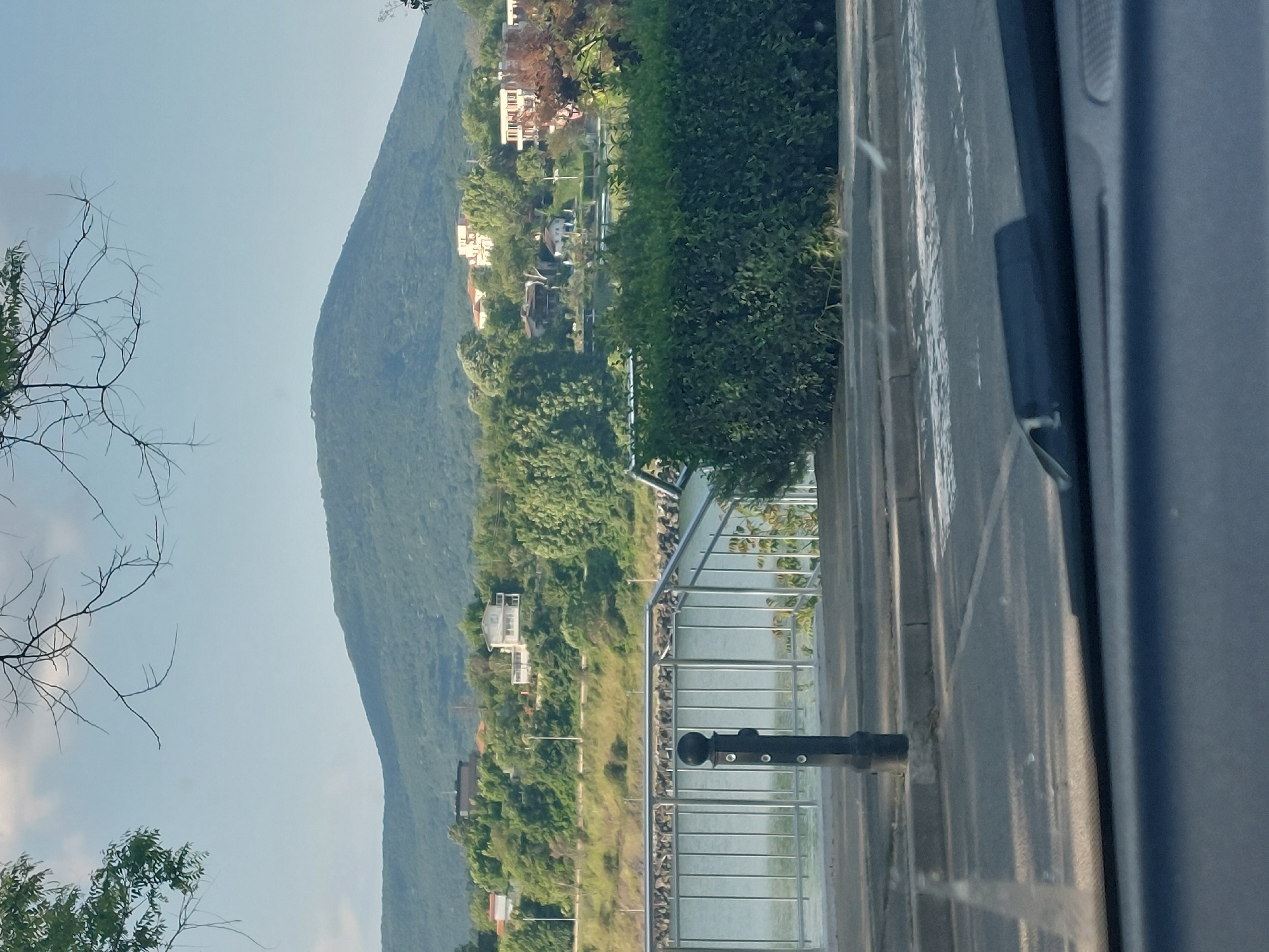 From an elevated position, possibly a vehicle on a road, a landscape is captured. A large, forested hill dominates the left side of the frame. Along the base of the hill are scattered houses and a road with vegetation. A metal railing and concrete barrier line the right side of the road, separating it from a body of water. Beyond the barrier, there's lush greenery and possibly some beach/river area.
Given the context of Tsarevo, Bulgaria, it is likely that this scene depicts the coastline or a river running through the area. The hill could be one of the small mountains around the city, and the body of water may be the Black Sea or a local river. There is not enough detail to state more definitively without further visual data.
