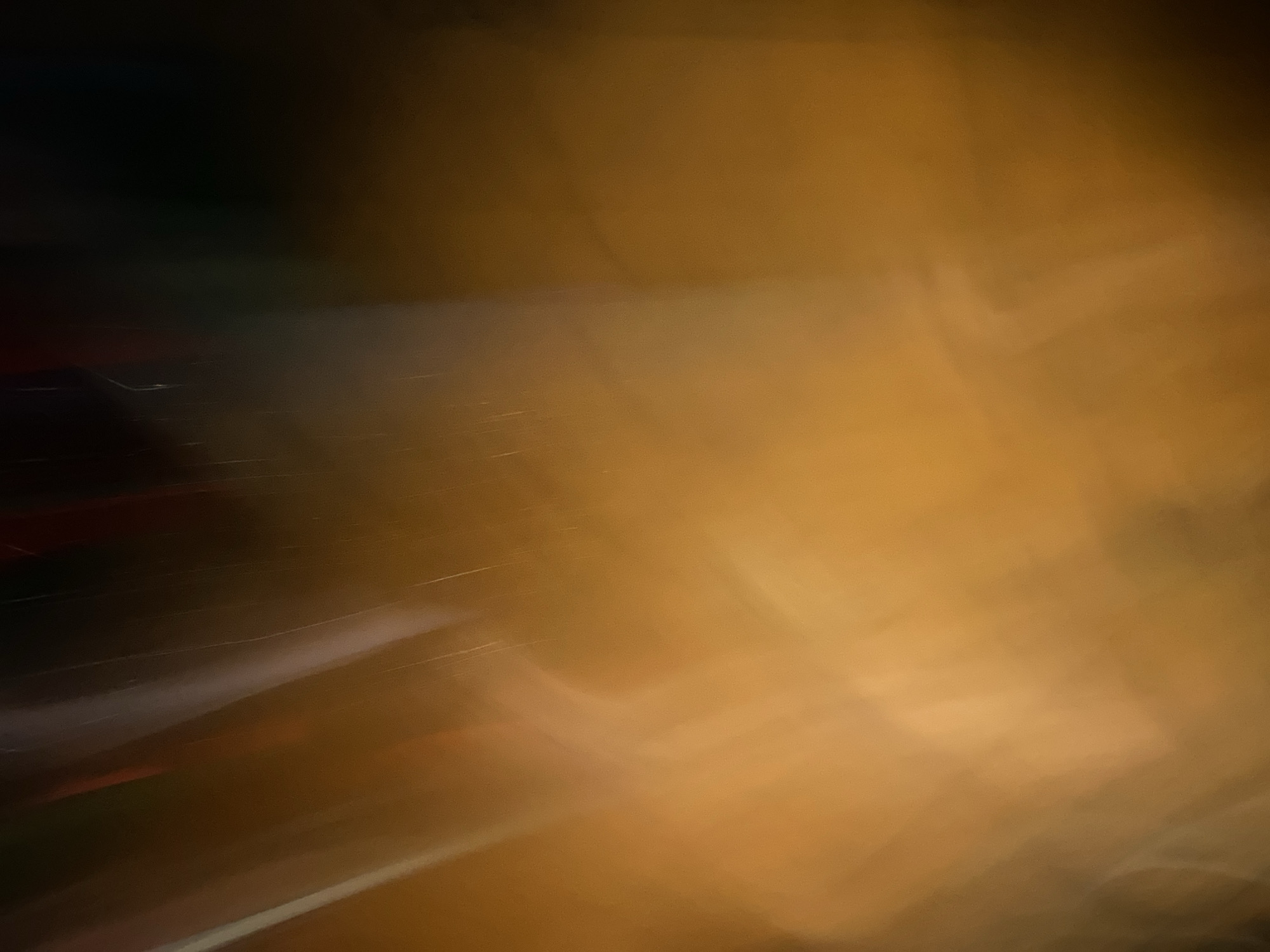 This abstract image presents a highly blurred and out-of-focus scene dominated by warm hues of amber and deep brown, transitioning from brighter, diffused light on the left to darker, more muted tones on the right and top. Diagonal streaks of diffused white and light gray, possibly indicative of moving light sources or reflections, are sparsely visible, particularly in the upper left quadrant. The overall impression suggests a light-streaked environment captured with significant motion blur or a shallow depth of field. There are no identifiable human subjects, specific objects, or discernible actions. The generalized environment is consistent with artificial lighting in an urban setting or interior space. This visual capture is situated in the city of Howayi, Nigeria.