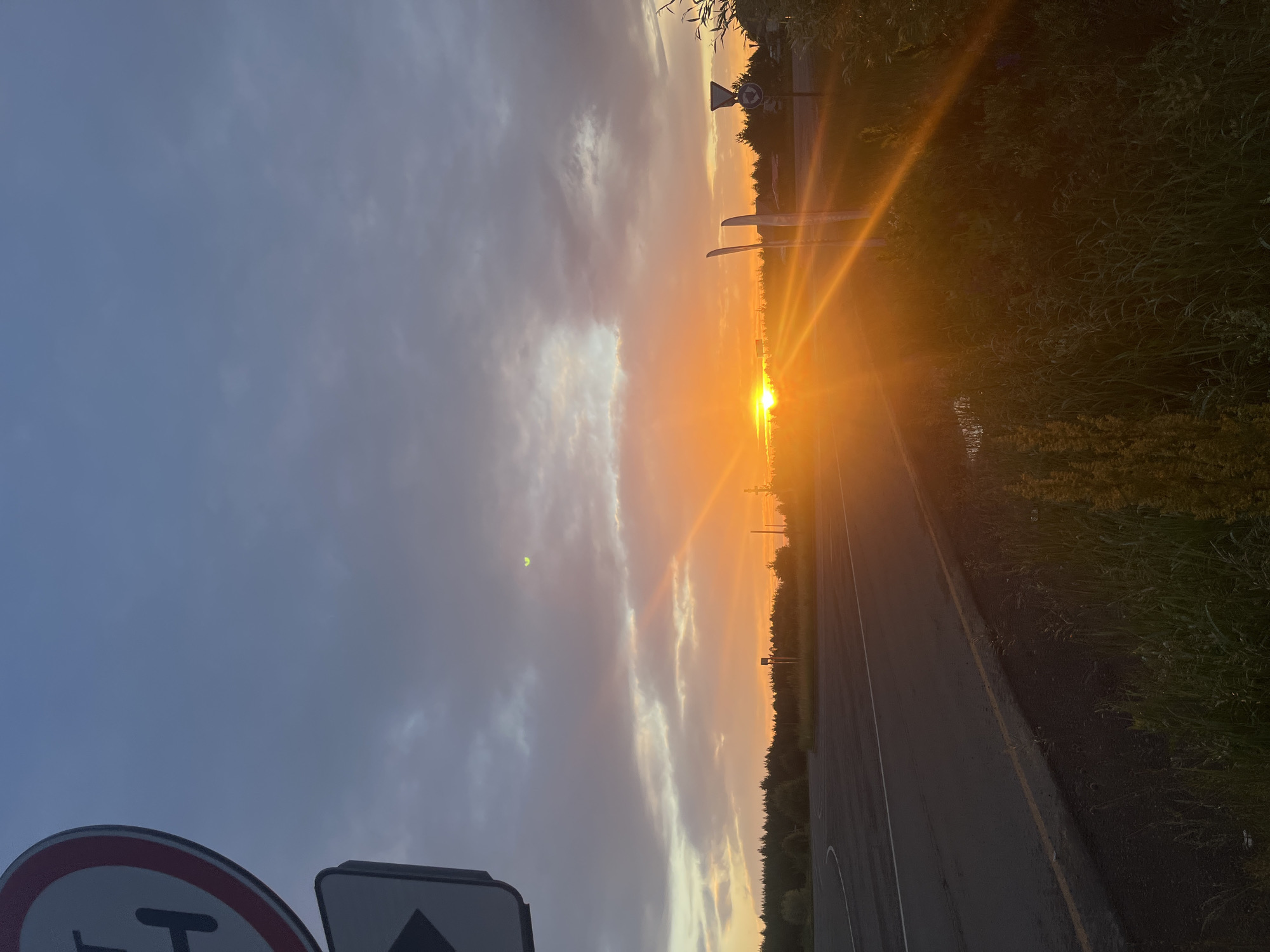 The image shows a sunset over a rural road in Gol'yany, Russia. The sky is filled with dramatic clouds, ranging from dark gray to bright orange and yellow, illuminated by the setting sun. The sun itself is a brilliant orb, casting long rays of light across the scene. A paved road curves into the distance on the left side of the image, with a yellow line marking the center. On the right side of the road, there is a patch of tall grass and foliage. Several road signs are visible, including a circular sign with a red border and a black "H" on a white background on the left, and a triangular sign with a red border and a black arrow on a white background below it. Further down the road, another circular sign with a red border and a blue background, indicating a roundabout, is present. Two tall, slender, white poles stand near the road, and the glint of sunlight reflects off their surfaces. The distant horizon is lined with a dense forest. The overall scene is one of natural beauty and the tranquility of a rural sunset.