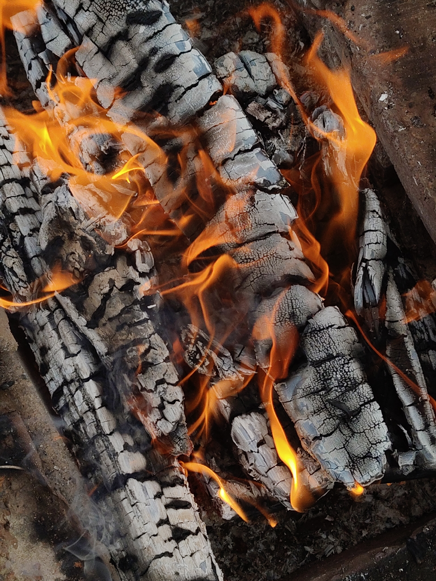 A close-up shot depicts a fire burning through charred wood. The wood is gray and fractured, with cracks and a rough texture. Bright orange and yellow flames engulf the wood, rising and swirling around the logs. The environment appears to be an outdoor fire pit or similar contained space. Location details are unavailable, so 'Unknown, Unknown' city information cannot be provided.