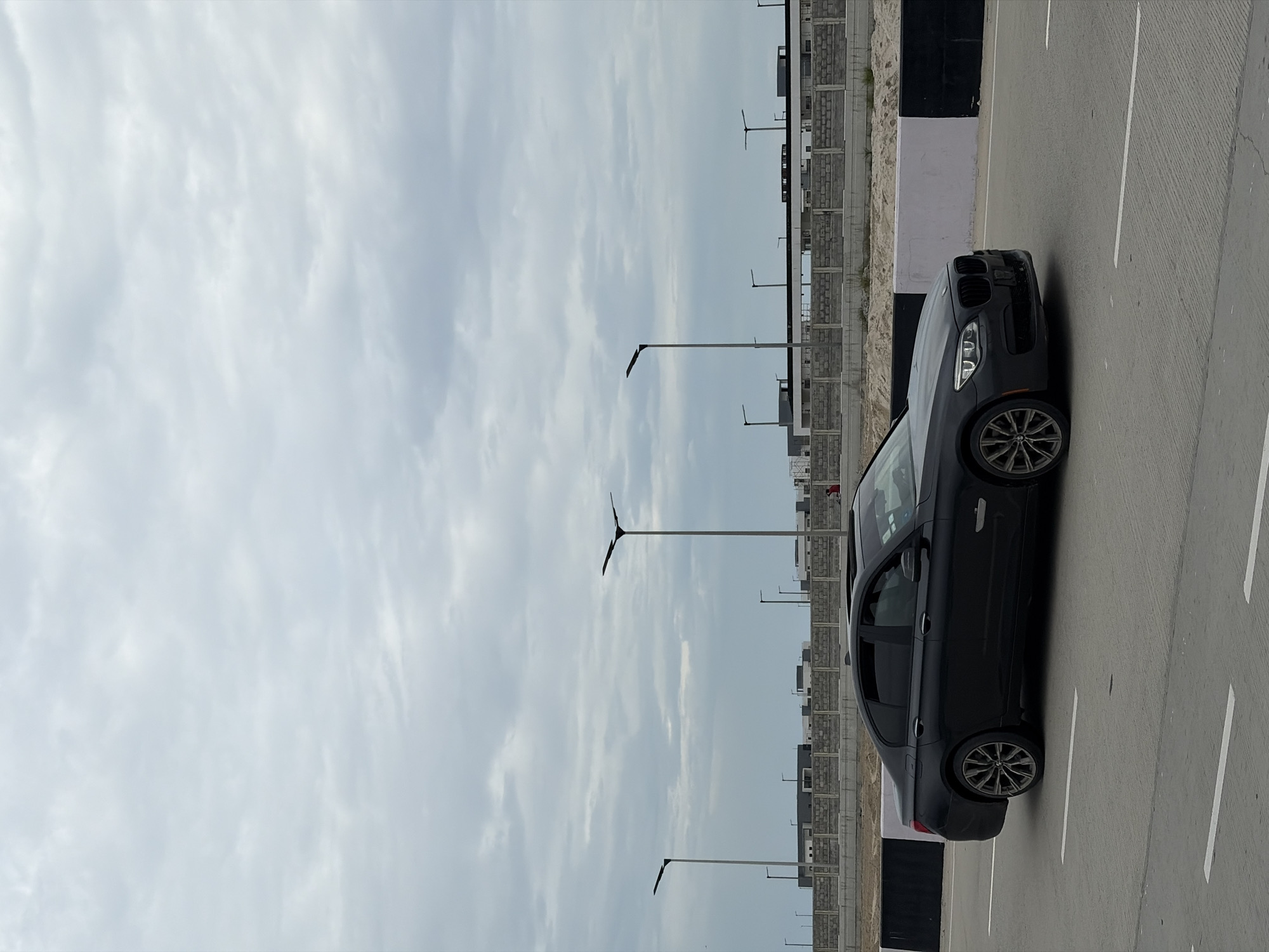 A black BMW sedan is captured in motion on a paved road in Igboefon, Nigeria. The car is positioned in the right half of the frame, with its front facing towards the right. The vehicle appears to be a four-door sedan with tinted windows and a sunroof. Its headlights are illuminated.
The road surface is asphalt, marked with white lane lines. Along the left side of the road, there is a construction site featuring sand, concrete barriers with black and white markings, and various streetlights. In the background, a low-rise building complex is visible, with a concrete wall in front of it. The sky is overcast with diffused clouds. A single person in a red shirt can be seen walking in the background near the construction site.