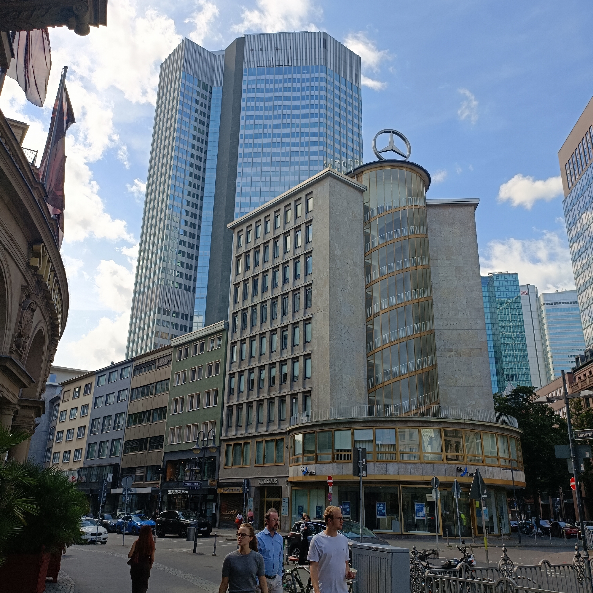 The image depicts a street scene in Frankfurt, Germany, focusing on the Eurotheum skyscraper and surrounding buildings. In the foreground, pedestrians are walking on the sidewalk. A woman with long auburn hair is walking away from the camera, wearing a dark dress. Two men and a woman are walking towards the camera. The man on the right is wearing a white t-shirt and glasses, holding a white cup. The man in the middle wears a blue collared shirt and glasses. The woman on the left wears a grey t-shirt and glasses. Several cars, including a white sedan, a blue sedan, and a black SUV, are parked or driving on the street. A motorcycle is parked on the sidewalk.
The environment includes buildings of varying architectural styles and heights. The prominent Eurotheum skyscraper, with its distinctive silver and blue facade, dominates the background. In front of it is a circular building with a Mercedes-Benz star logo at its apex, featuring a glass facade with balconies. To the left of the Eurotheum are several mid-rise buildings with a more uniform, rectangular design, some painted in muted colors like grey and green.
On the street, there are traffic signs, street lamps, and planters with palm trees. The sky is partly cloudy with a clear blue sky visible. The scene appears to be a commercial district, with storefronts visible on the lower levels of some buildings, including "JUNIORHAUS," "BB Bank," and "Shoepassion." The street is paved with cobblestones in some areas. The overall impression is of a busy urban environment.
