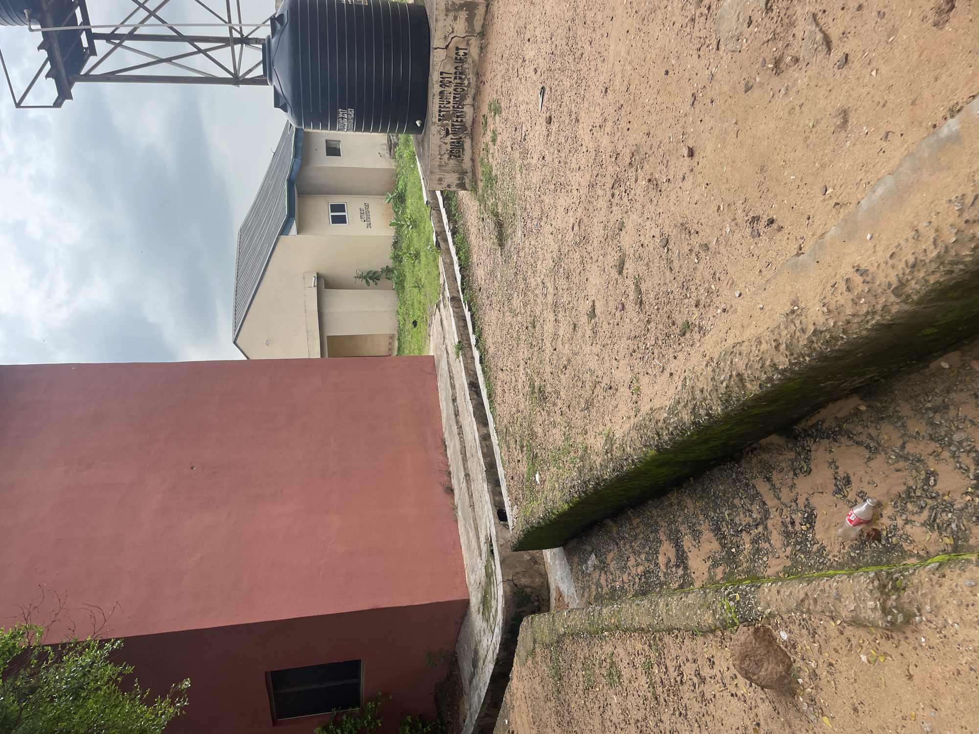 In Lokoja, Nigeria, the image captures an outdoor scene dominated by construction and infrastructure elements. A large, red-walled building occupies the left side of the frame, featuring a dark-paned window. In the background, a light-colored building with a metal roof stands adjacent to a tall, dark water tank supported by a metal structure. The ground is a mixture of dry earth and sand, with a concrete drainage channel running through the foreground. This channel appears somewhat overgrown with moss and contains some debris, including a discarded plastic bottle. The sky is overcast with clouds. The scene is static and devoid of human presence, focusing on architectural features and the surrounding, somewhat neglected, environment.