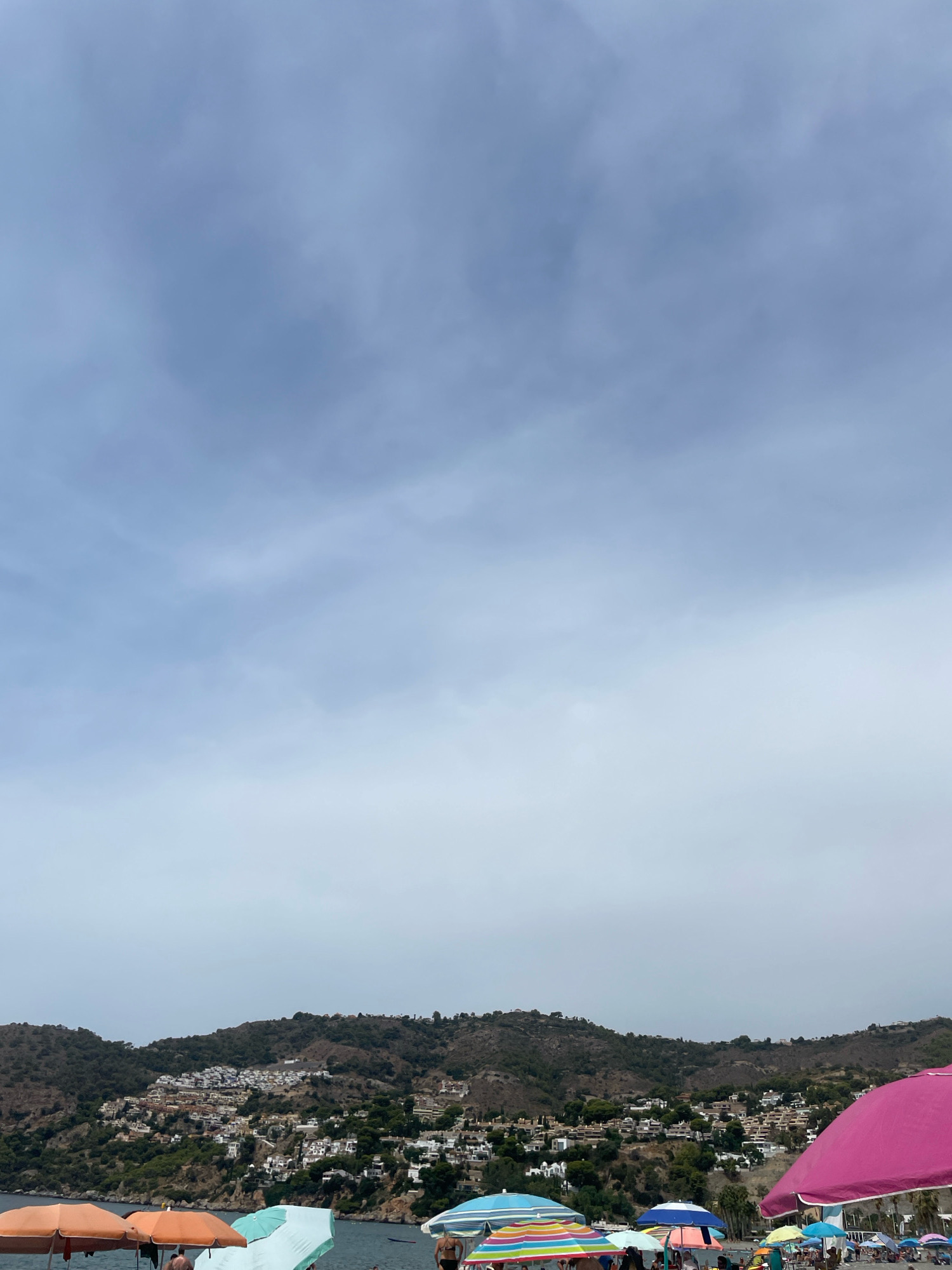 The image shows a crowded beach in La Herradura, Spain, with a view of the town built on a hillside in the background. Numerous beach umbrellas, in various colors including pink, orange, blue, and striped patterns, are set up on the sandy shore. Some people are visible under the umbrellas and walking on the beach. The sea is calm and blue. The sky is overcast with light clouds.
