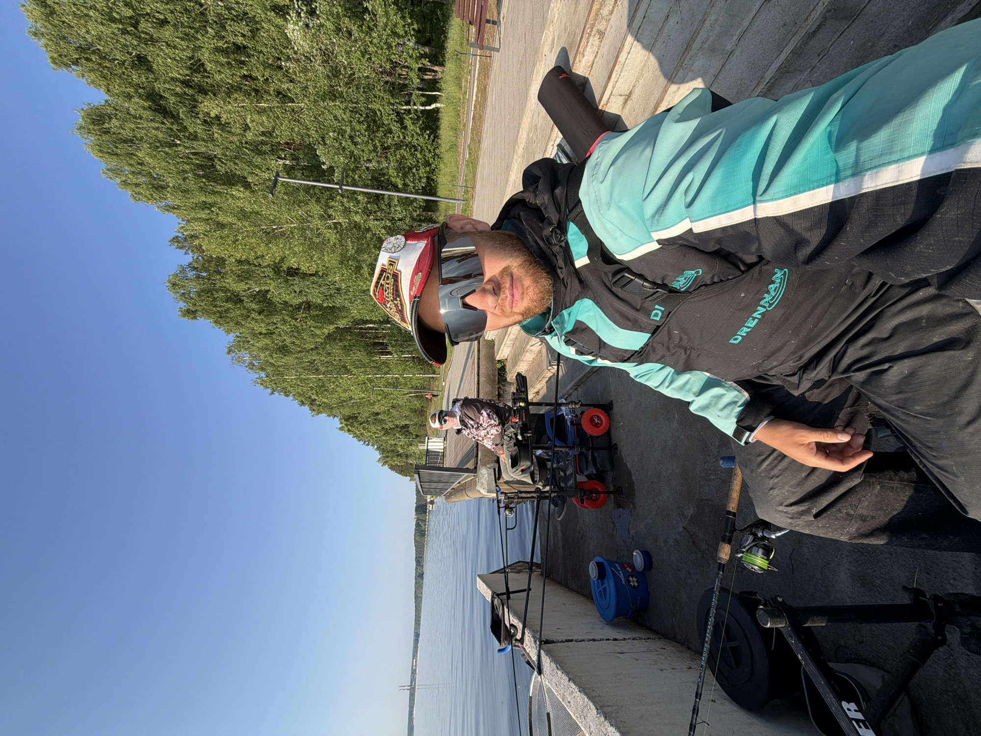 The image depicts a sunny day scene by a body of water, likely a reservoir, in the city of Chaikovsky, Russia. In the foreground, a man wearing a fishing vest and a baseball cap is seated in a fishing chair. He is holding a fishing rod and looking at the camera. Beside him, another person is also seated in a fishing chair with a fishing rod. The background features a line of birch trees and a clear blue sky. The setting appears to be a pier or embankment, with fishing equipment such as rods, a net, and buckets visible. The overall atmosphere is one of leisure and outdoor activity.