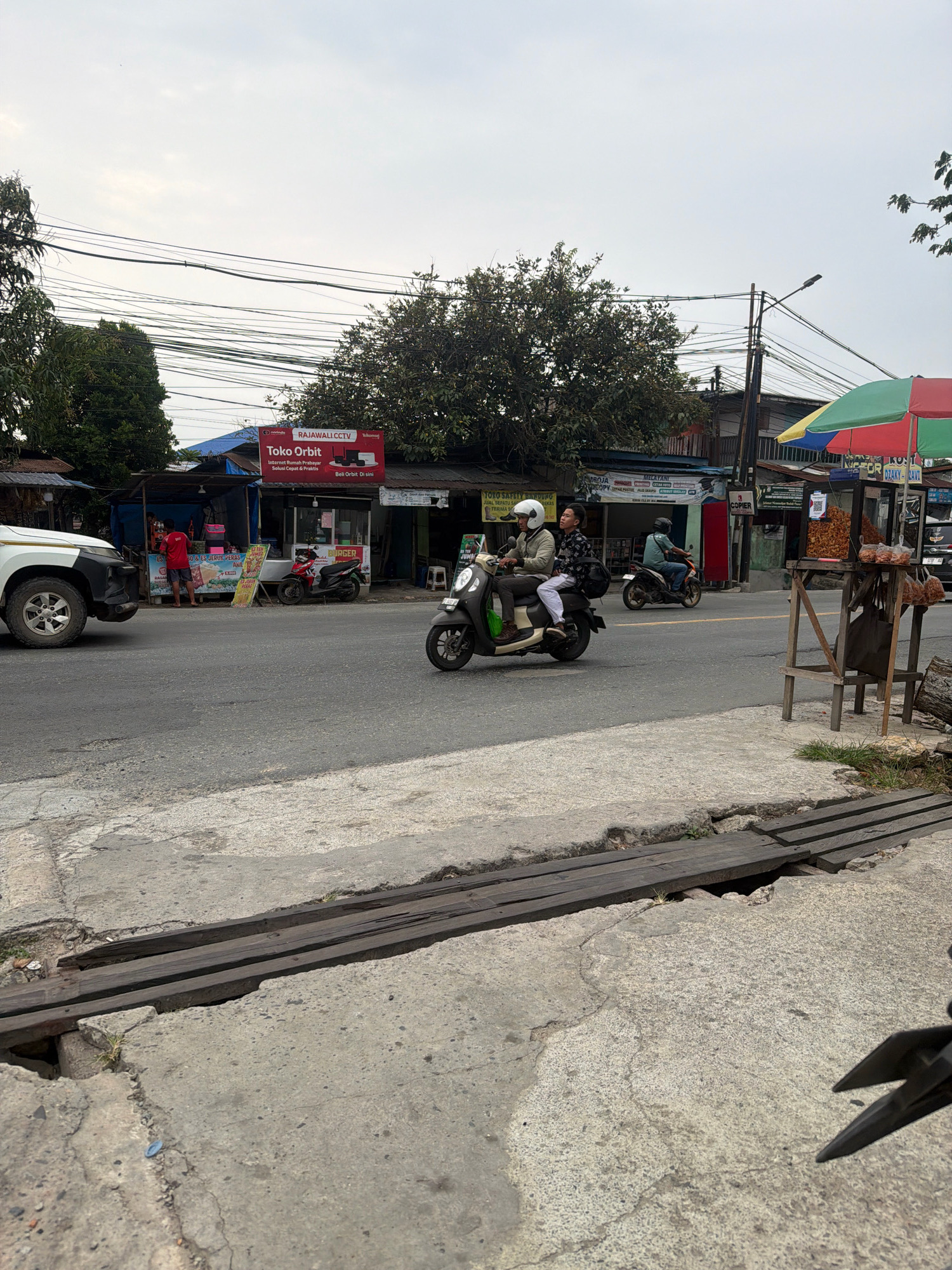 The image depicts a street scene in Balikpapan, Indonesia, under an overcast sky. A grey asphalt road occupies the foreground and midground. On the road, two individuals are riding a dark-colored scooter, with the driver wearing a white helmet and a patterned shirt, and the passenger wearing a white helmet and a grey jacket. To the left, a white SUV is partially visible, parked on the side of the road. In the background, a row of small businesses with corrugated metal roofs line the street. One prominent sign reads "Toko Orbit," advertising internet services and CCTV. Colorful umbrellas, red, green, and yellow, are visible near a stall selling fried snacks. A wooden stand with stacked snacks and bags is positioned near the right edge of the frame. Overhead, a dense network of power lines crisscrosses the sky. The sidewalk area in the foreground shows a damaged concrete surface with wooden planks covering a gap or hole.